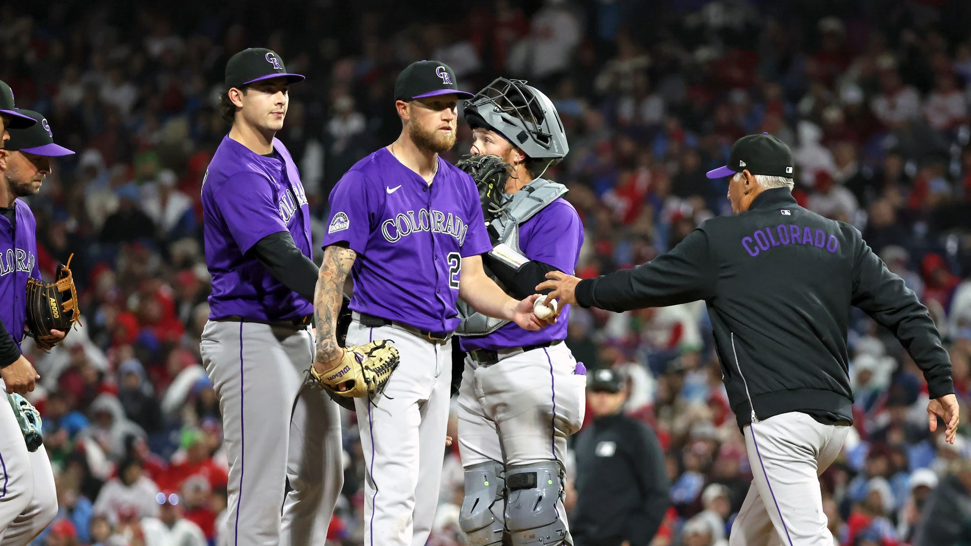 Baseball players stand on a mound, with one player handing over a baseball to a man in a black jacket. 
