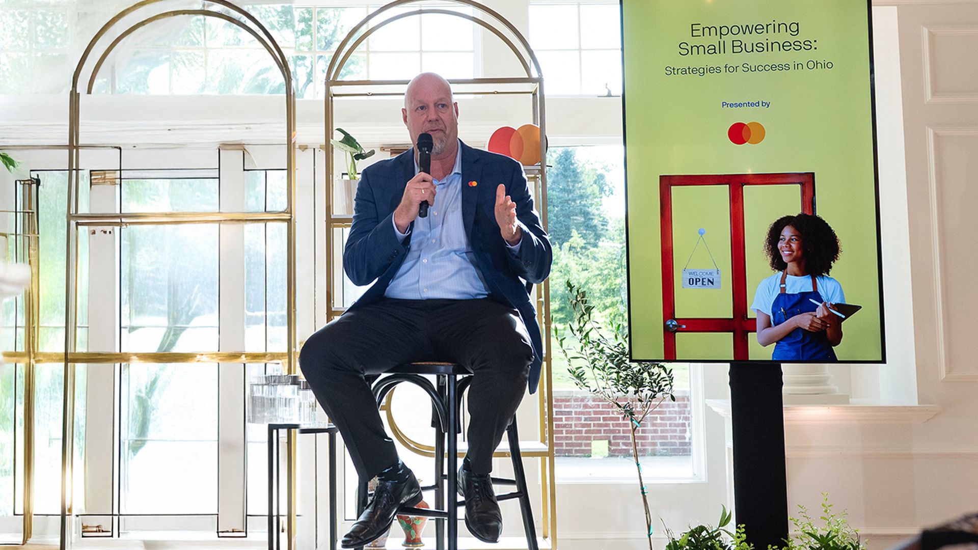 Man in a blue suit speaking into a microphone while seated, next to a screen with a presentation titled "Empowering Small Business: Strategies for Success in Ohio" featuring a woman in an apron.