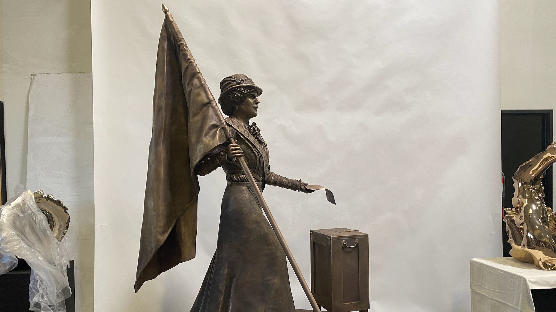 A side view of a bronze statue of a woman holding a flagpole in her right hand and a piece of paper in her left hand.