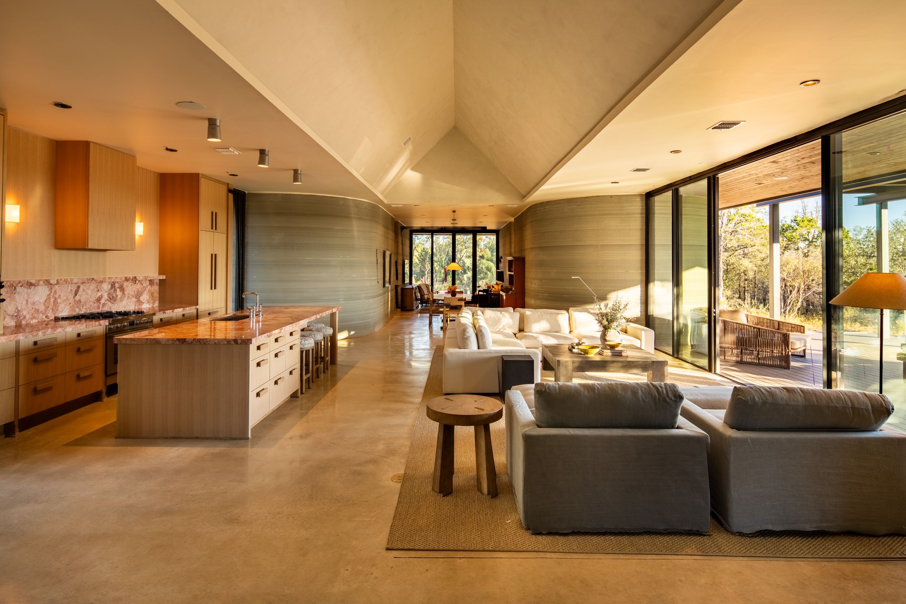 Modern open living space with beige floors, large cream sectional sofa, wooden coffee table, kitchen island with marble countertop, and floor-to-ceiling windows showing outdoor seating and greenery.
