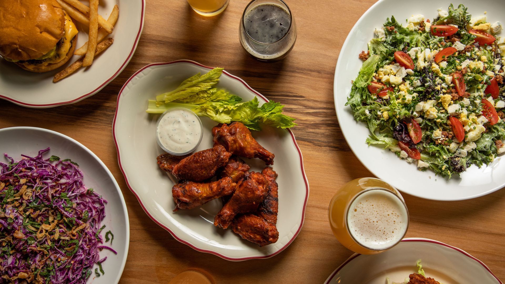 A full wooden tabletop covered in salads, chicken wings and beers is viewed from above. 