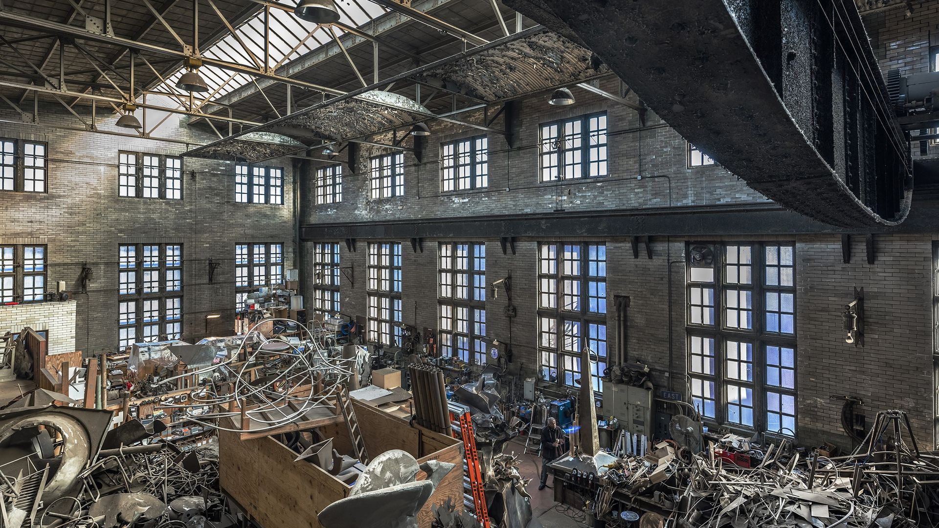 A large warehouse with windows and exposed beams with metal and wood scattered on the floor.