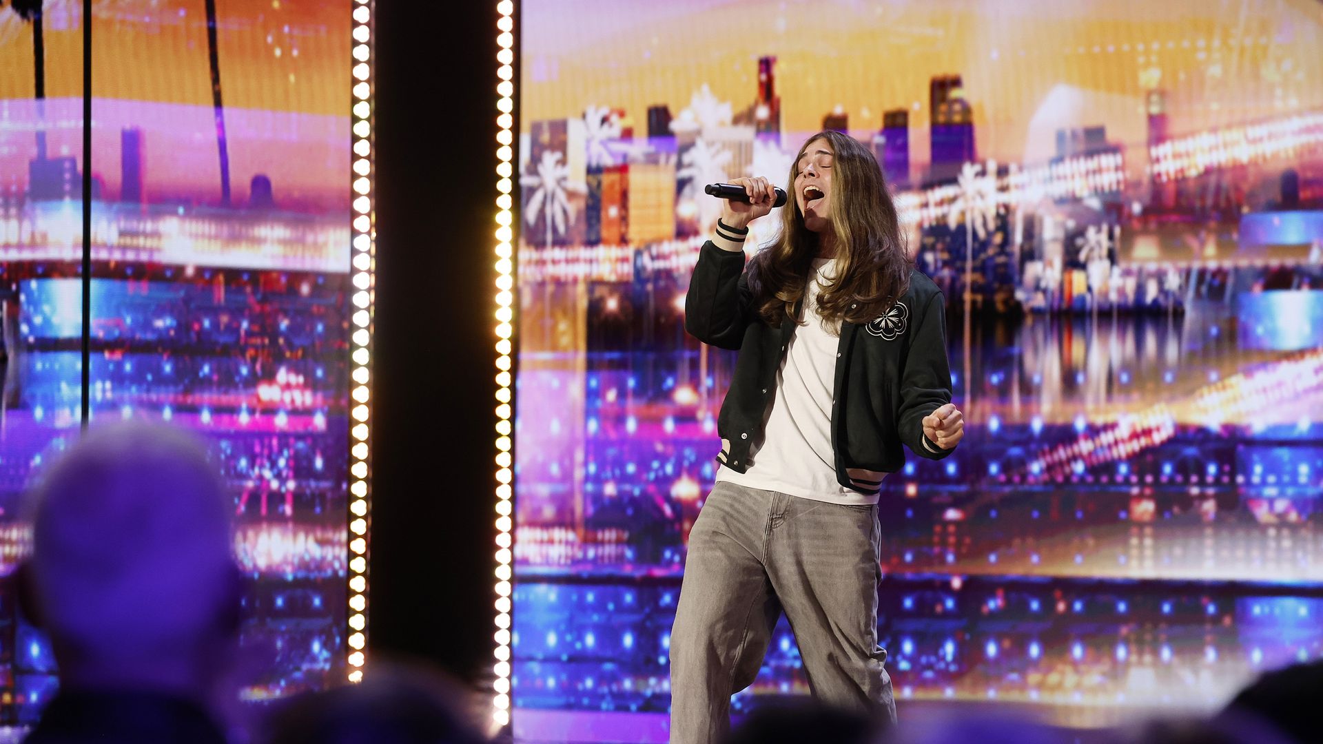 Young man with long brown hair singing passionately into a microphone on stage, wearing a black jacket, white shirt, and gray pants, with colorful cityscape lights in the background.