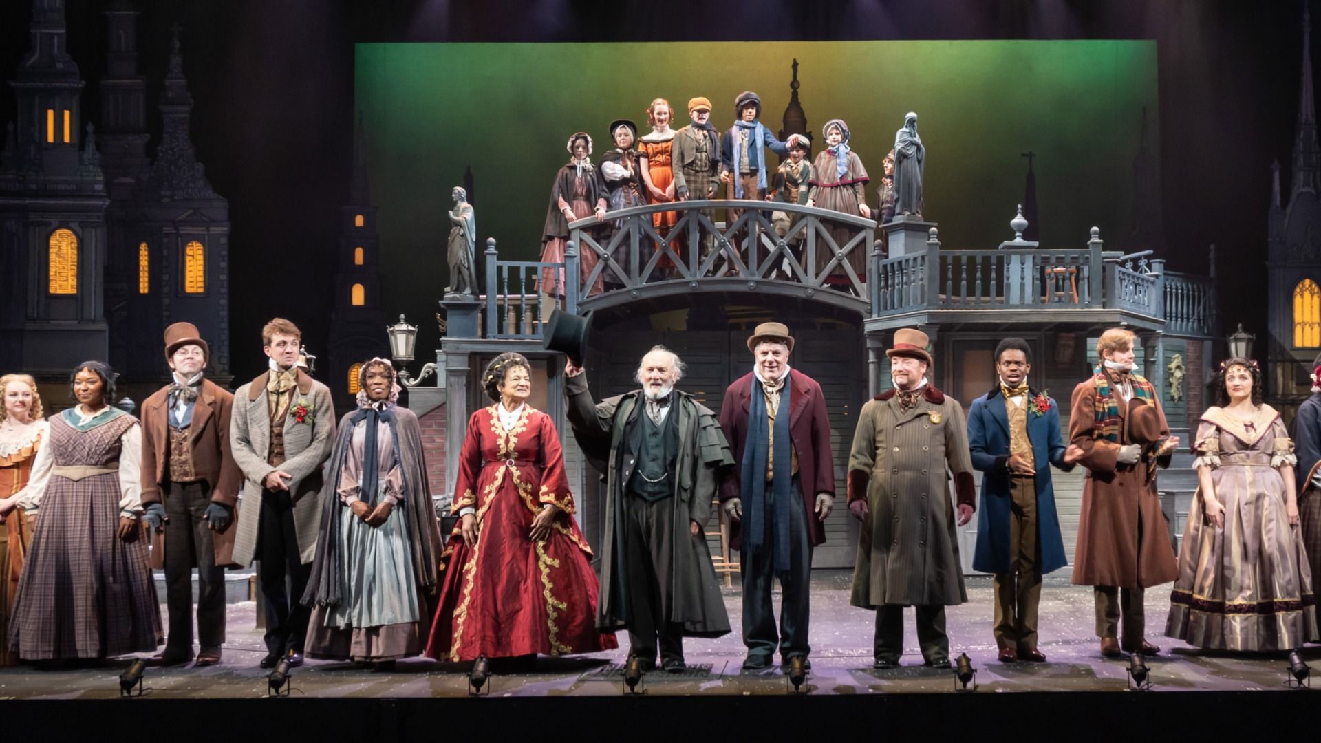 Actors in Victorian-era costumes take a bow on stage in front of a set with a bridge and lit windows, depicting a theatrical production.