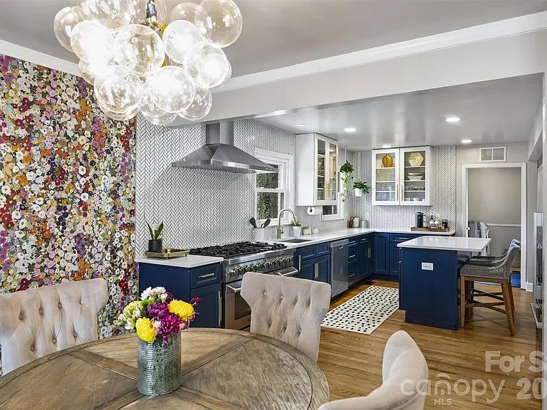 Bright kitchen with navy blue lower cabinets, white upper cabinets and countertops, floral accent wall, wooden round dining table with tufted beige chairs, and a glass bubble chandelier.