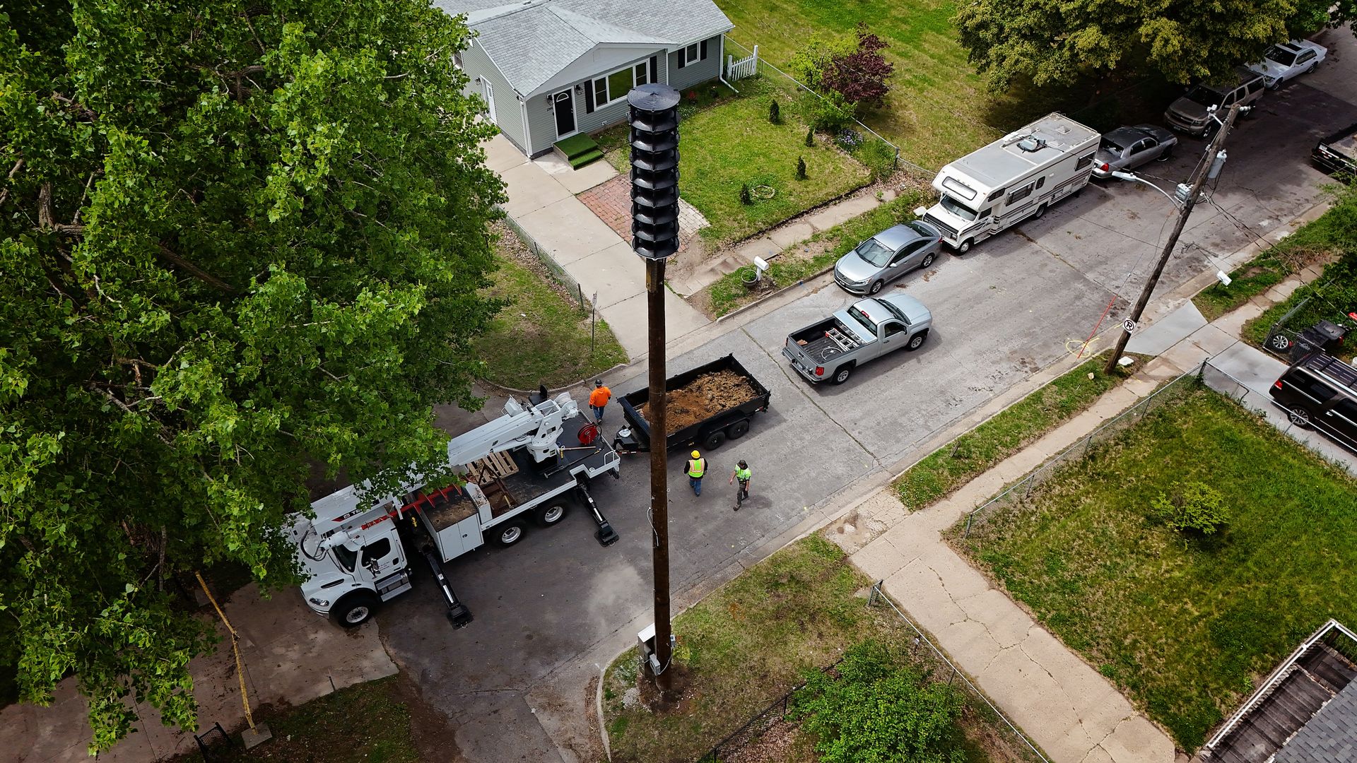 A tall outdoor warning siren tower in a Des Moines residential neighborhood with workers in safety vests near utility trucks on a street with houses, cars, and green trees and lawns in 2025.