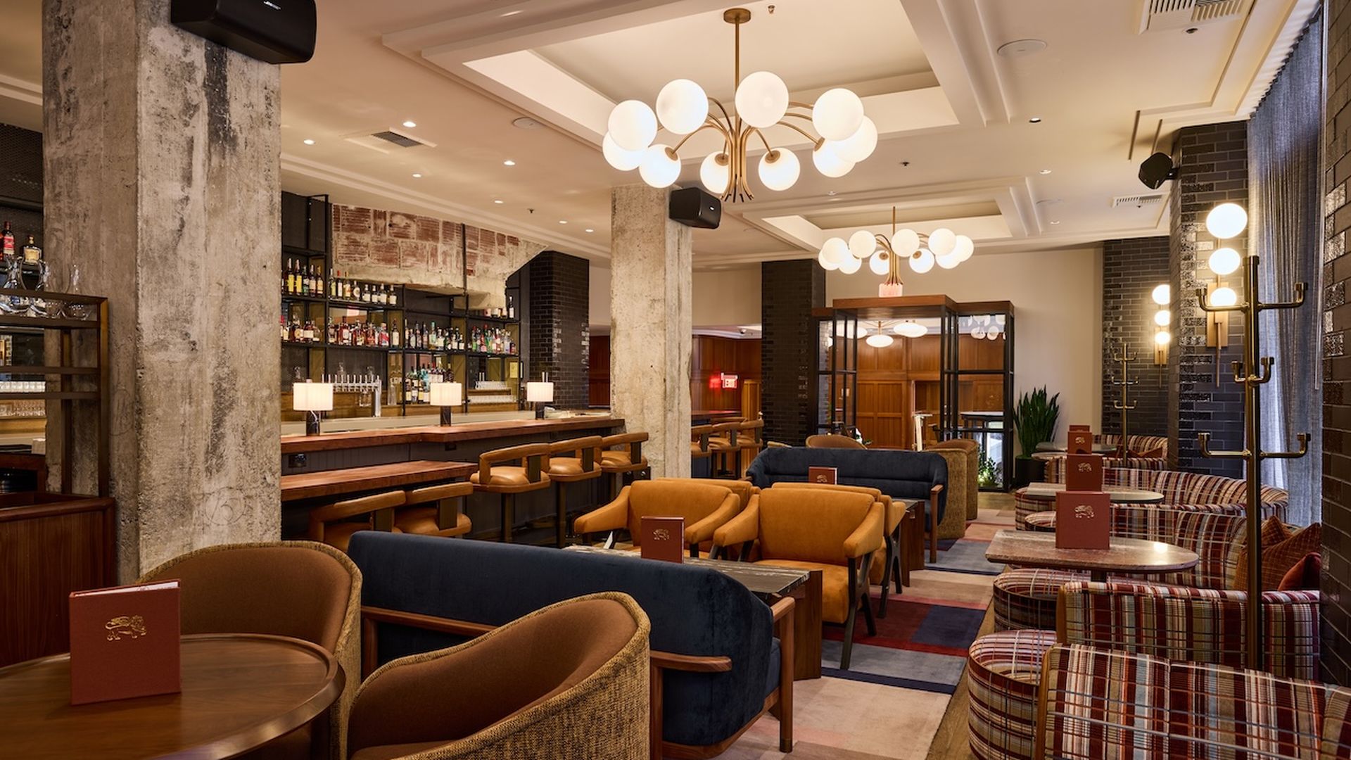 A stylish bar lounge with a long wooden bar, concrete columns, and plush seating. Round globe chandeliers illuminate plaid and beige chairs for a warm, modern ambiance.