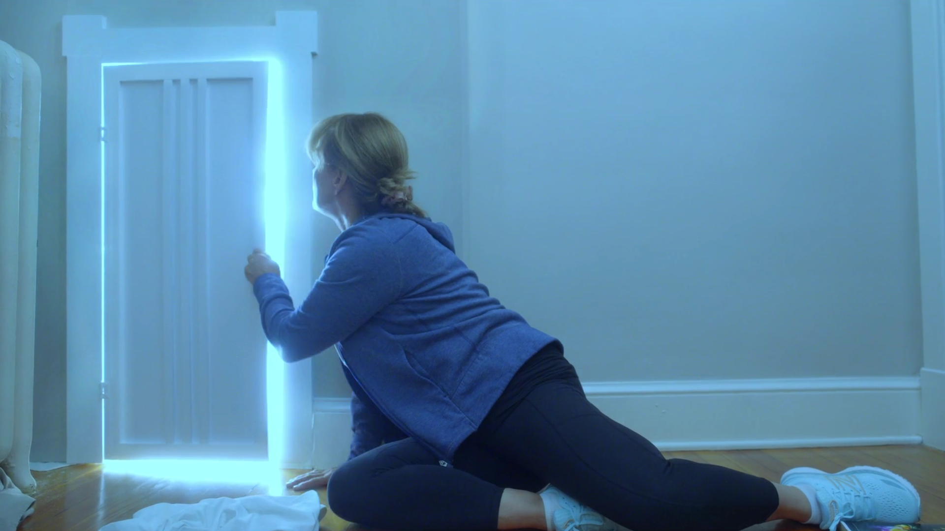 Person in a blue hoodie kneels on a wooden floor, facing a bright vertical blue light by a white door frame in a blue-tinted room; white sneakers lie nearby.