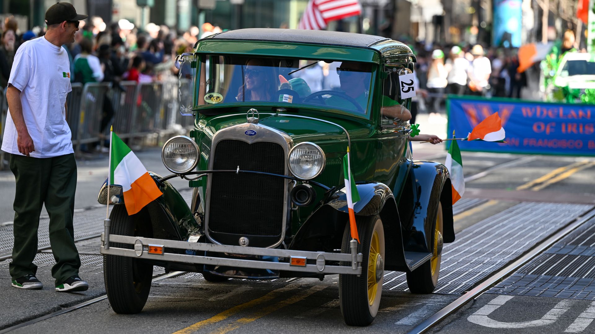 Vintage green Ford in a city parade, Irish flags on the bumper. Crowds behind barriers watch as a man in a white shirt stands to the left; banners and flags fill the background.