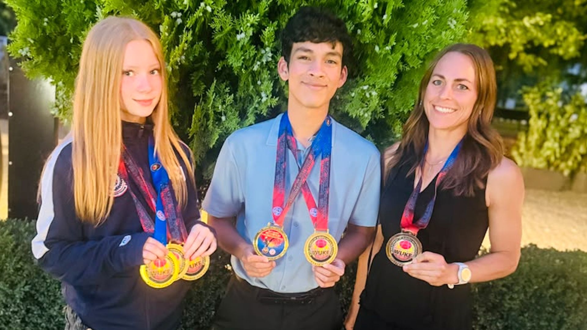 Three young people outdoors before a green hedge, proudly wearing gold medals on red and blue ribbons: a blonde girl in a navy jacket, a smiling boy in a light blue shirt, and a woman in black.