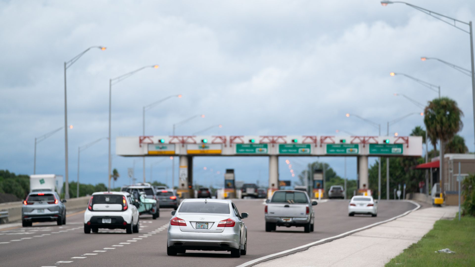 Multiple cars driving toward a toll plaza under a cloudy sky, with green lights showing open lanes and palm trees on the right side of the road.