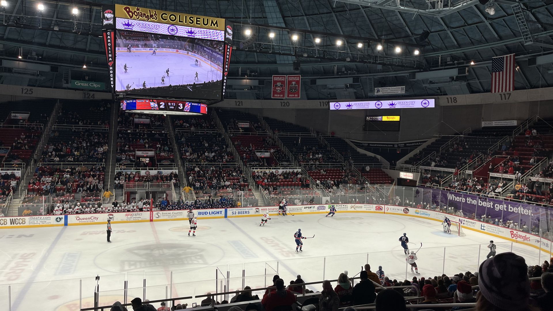 Indoor ice hockey arena at Bojangles' Coliseum with a large video scoreboard displaying a game, players on the ice, surrounding seats and banners, and an American flag overhead.