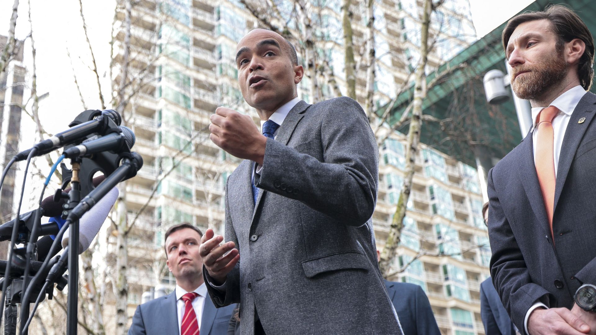 Nick Brown speaks with his hand slightly raised in front of a set of microphones, with two other men in suits at his right and left, with tall downtown buildings in the background.