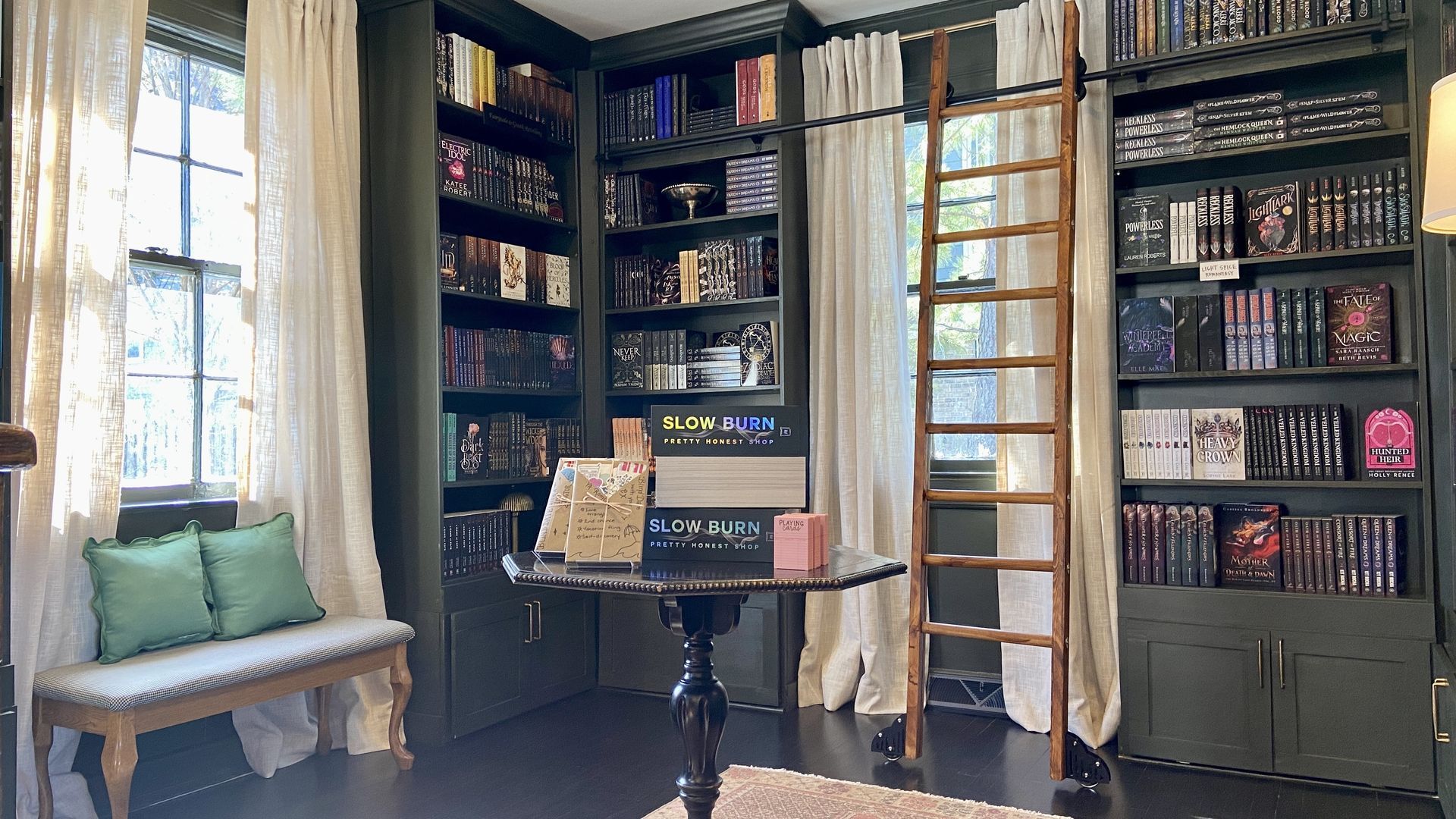 Inside Trope Bookshop. The dark green space is lined with bookshelves and books. There's a wooden ladder to the right in front of a window and a window seat to the left in front of another window. The floor-to-ceiling curtains are cream. An antique table holds books in the center of the photo.  