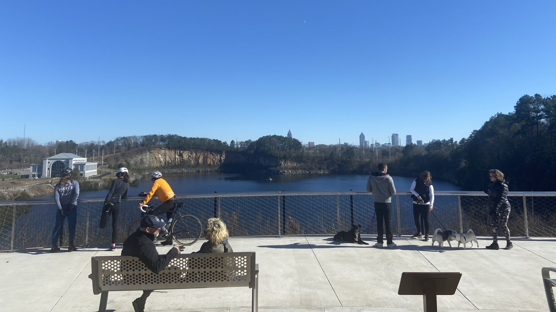 A group of bicyclists and walkers stand along a fence overlooking a reservoir made from a quarry with the city skyline in the background