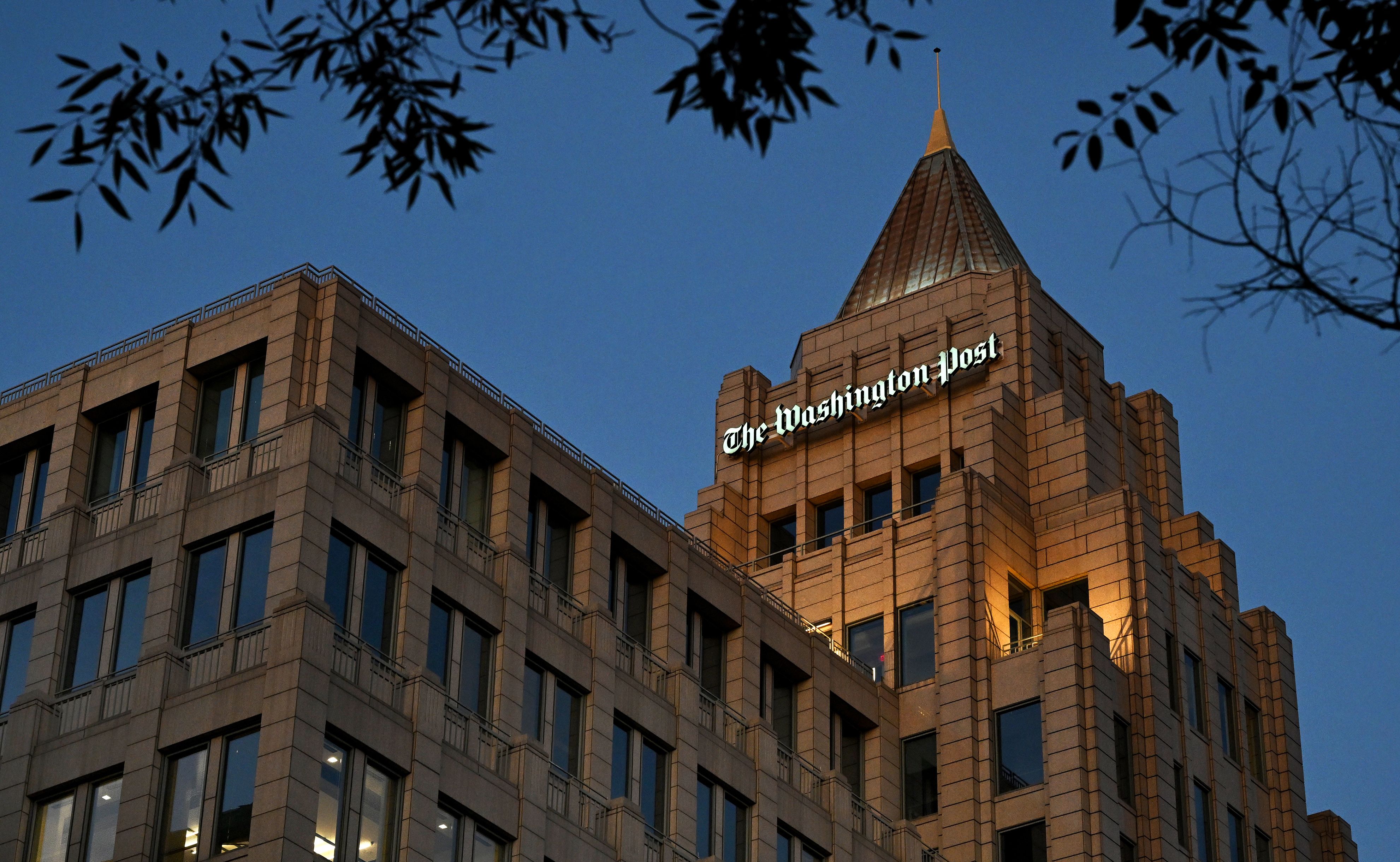 Evening view of The Washington Post building with illuminated sign on tan brick facade against deep blue sky, framed by dark tree branches at top corners.