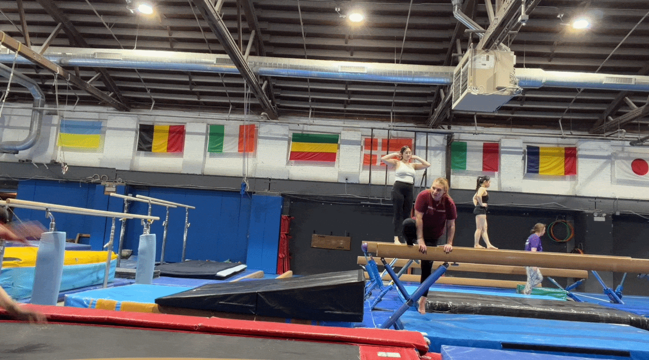 Gymnastics gym scene with a balance beam in the foreground; a coach kneels on the beam while a student and others stand nearby. Bright mats, blue walls, and international flags along the back.