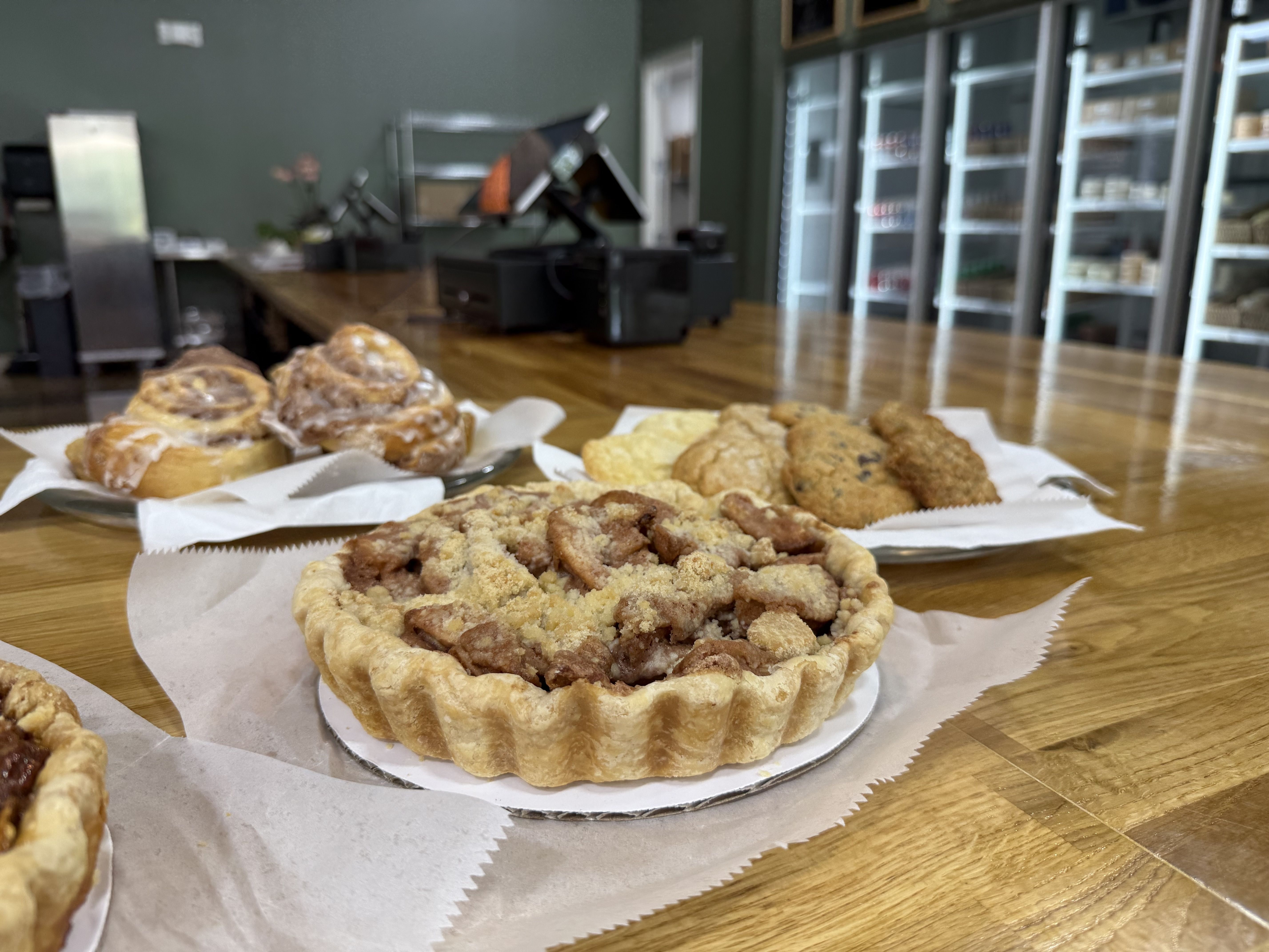 Close-up of a pecan pie with crumb topping, cinnamon rolls with icing, and assorted cookies on plates atop a wooden counter in a bakery with refrigerated shelves in the background.