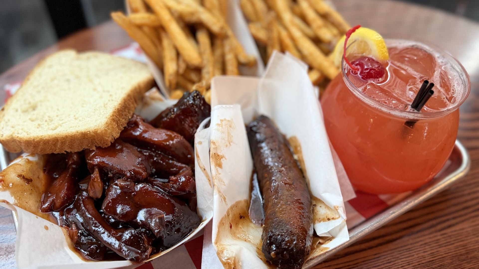 Tray of barbecue with glossy ribs, cornbread slice, sausage, fries, and a pink citrus cocktail with lemon wedge on a checkered tray.