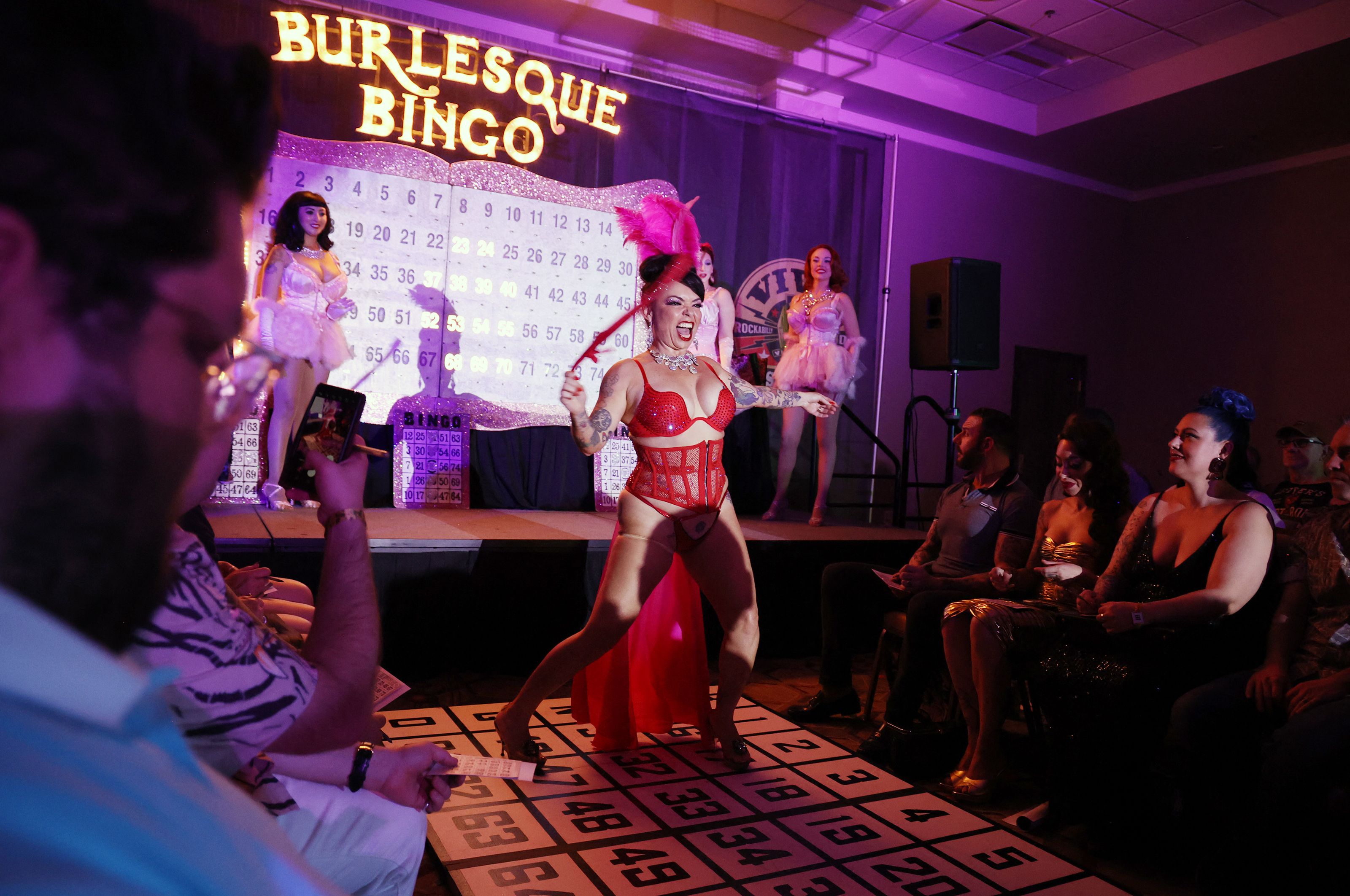 Costumed performers on a stage dance in front of a giant bingo card.