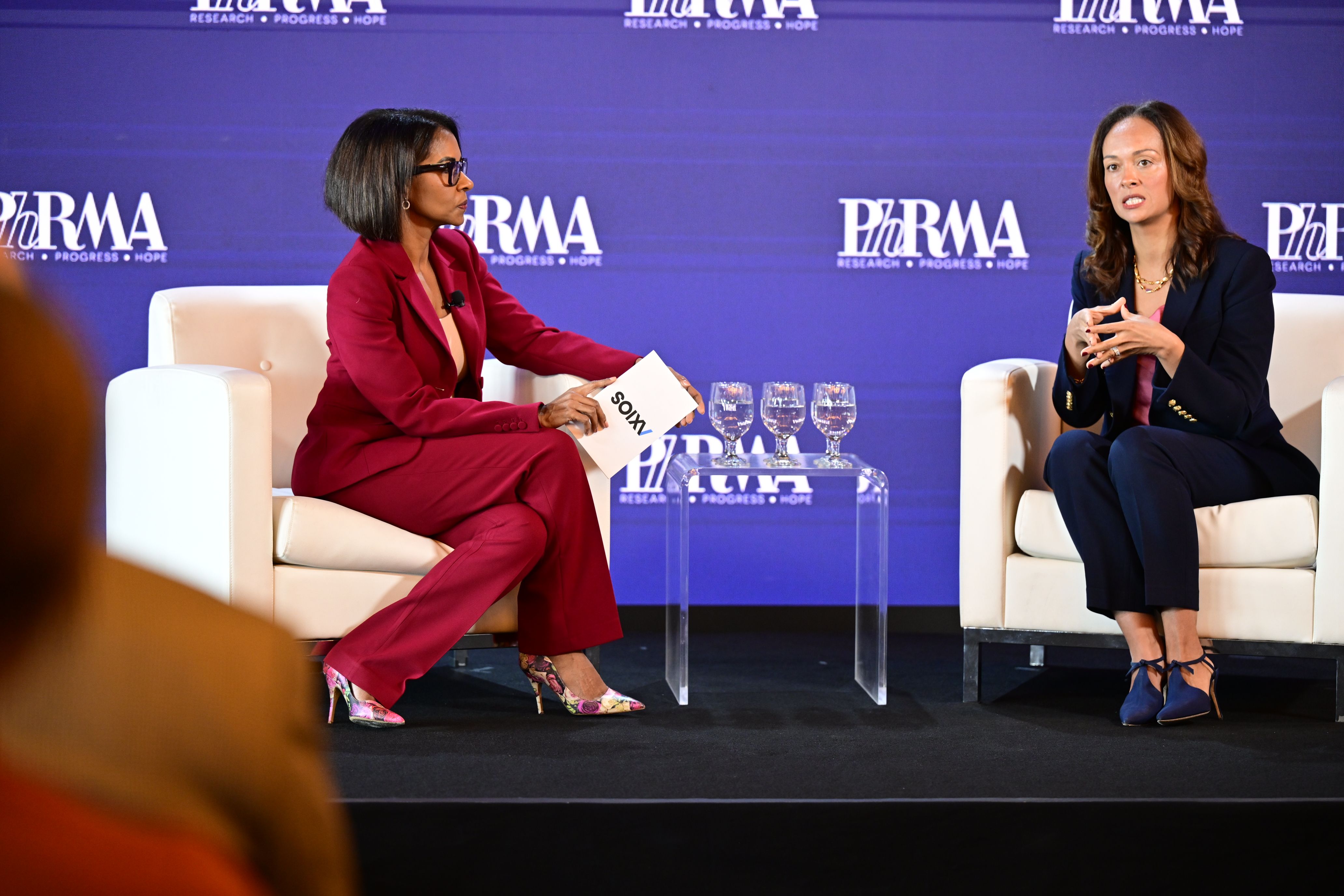 Two women in business attire on a purple PhRMA stage converse; left woman in red holds a card, right in navy gestures. A glass table with water sits between them.