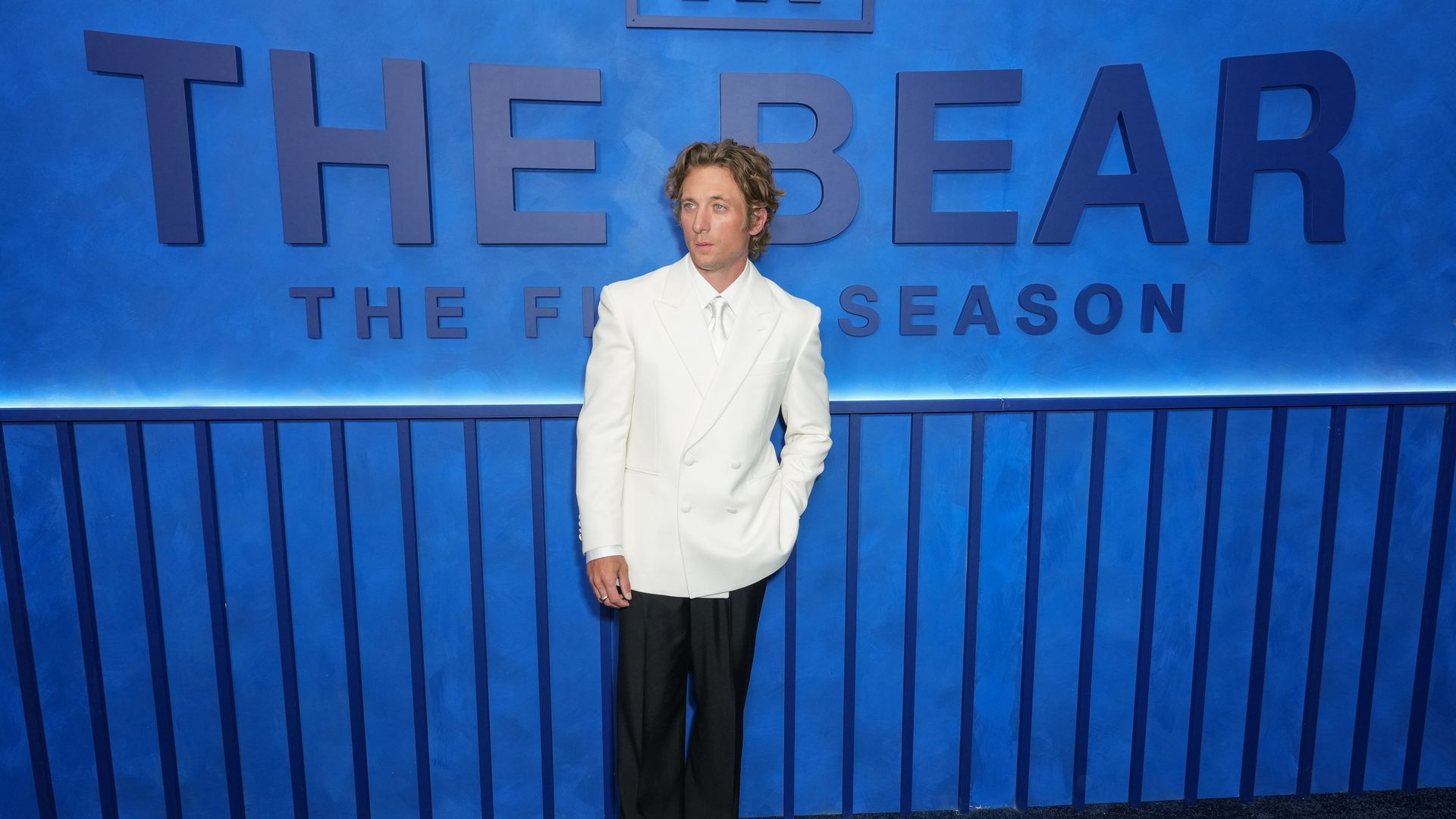 Man in a white suit stands against a blue wall with oversized letters spelling 'THE BEAR' and a small framed 'FX' logo above; he looks to the left with hands at his sides.