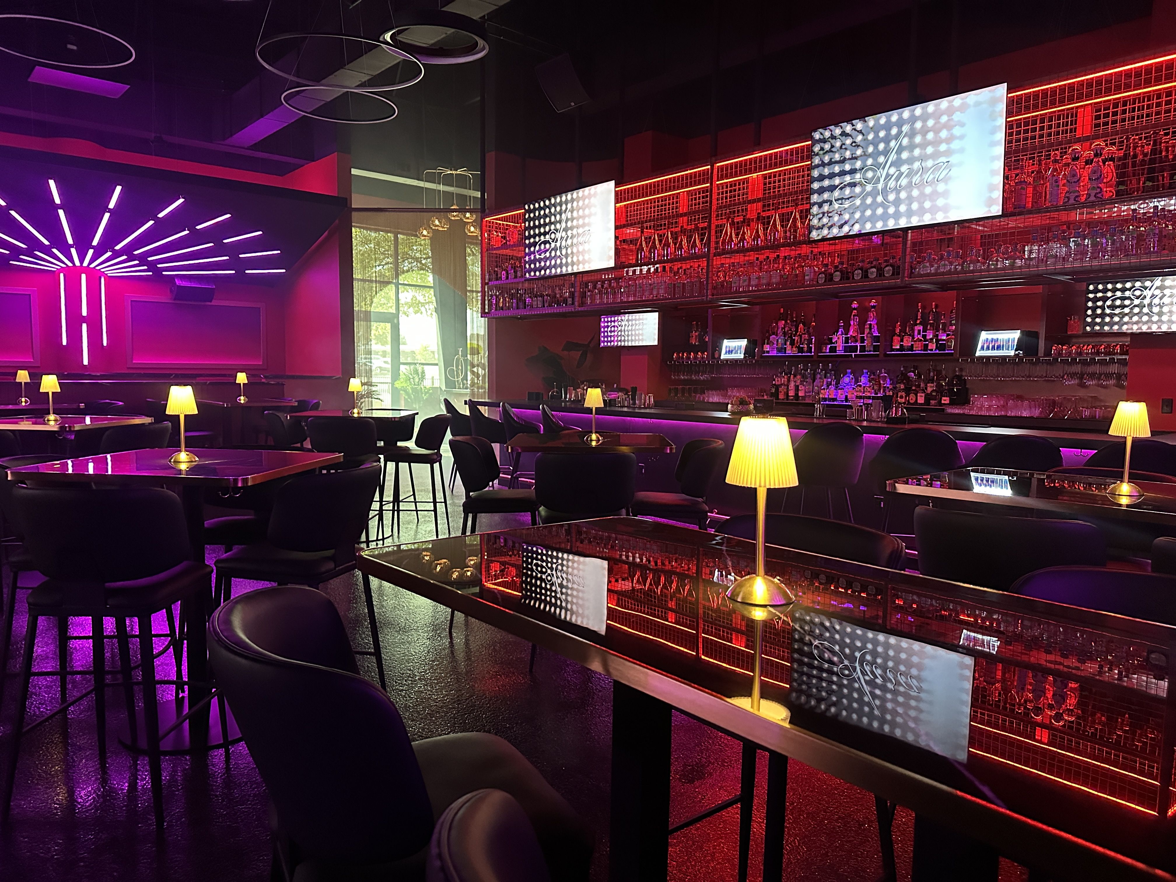 Modern bar interior with red and purple neon lighting, black chairs, and glossy tables topped with small yellow lamps. Screens display the name "Aura" above the bar.