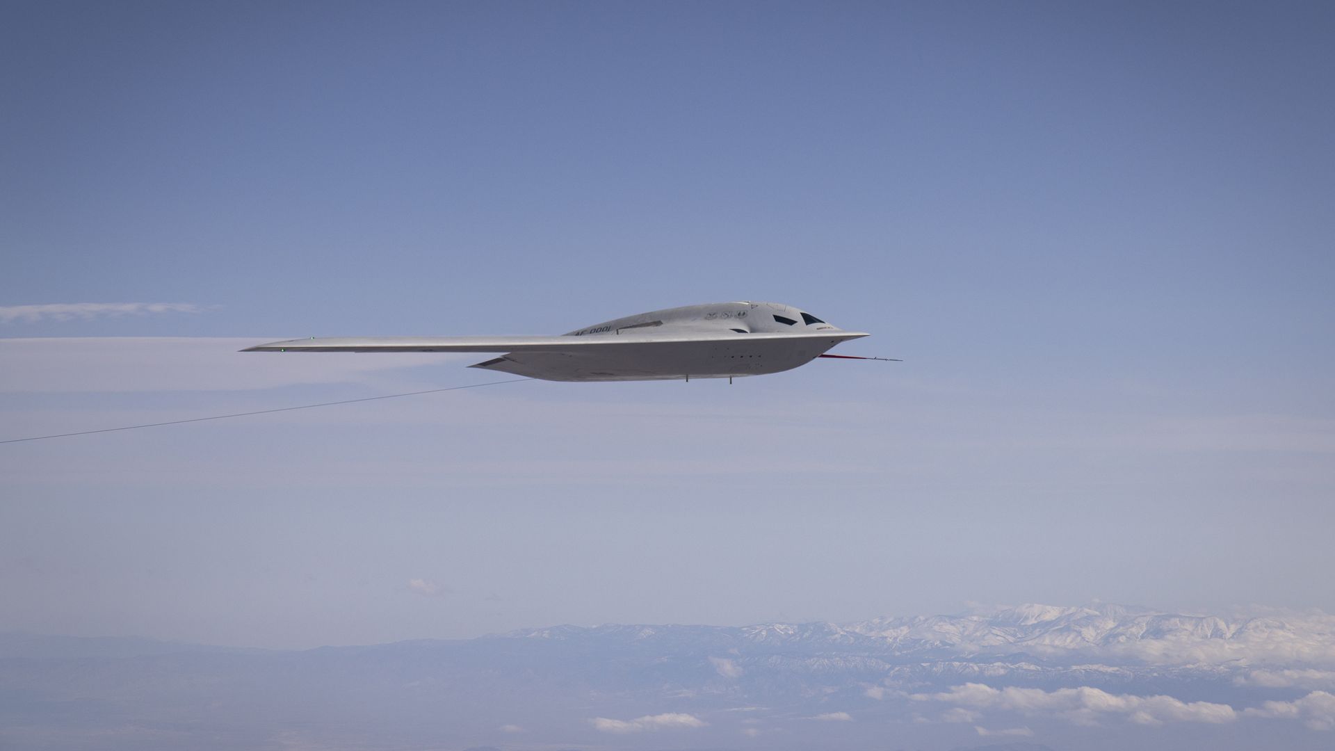 A sleek B-21 stealth bomber is seen flying above white clouds and against blue skies.