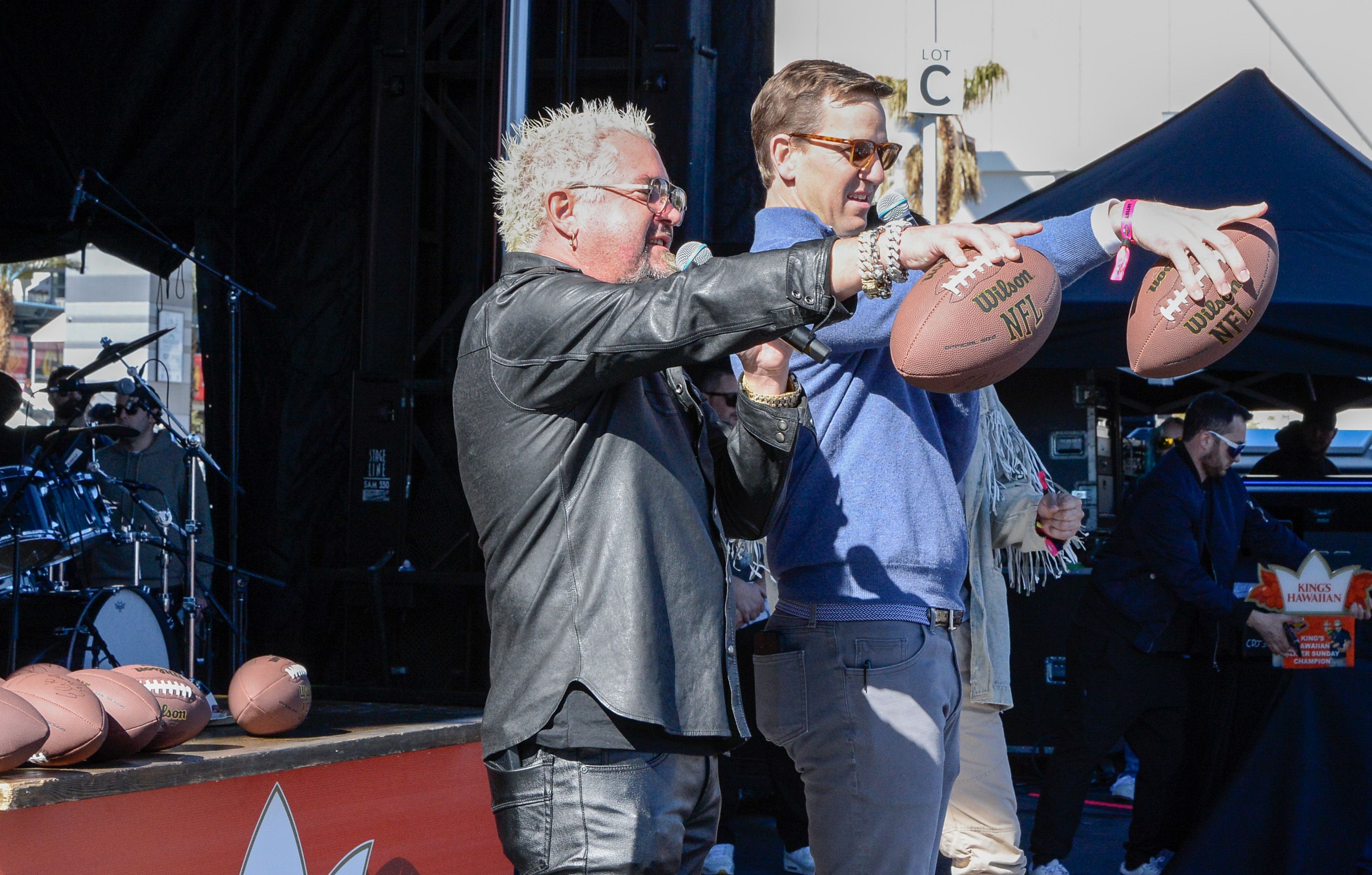 Guy Fieri and Eli Manning, both holding footballs in one hand and microphones in the other, gesture toward a crowd.