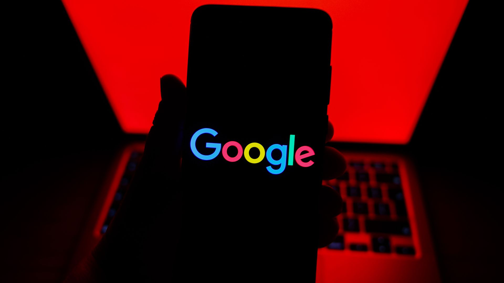 Hand holding a smartphone displaying the colorful Google logo in front of a red-lit laptop keyboard and screen in a dark setting.