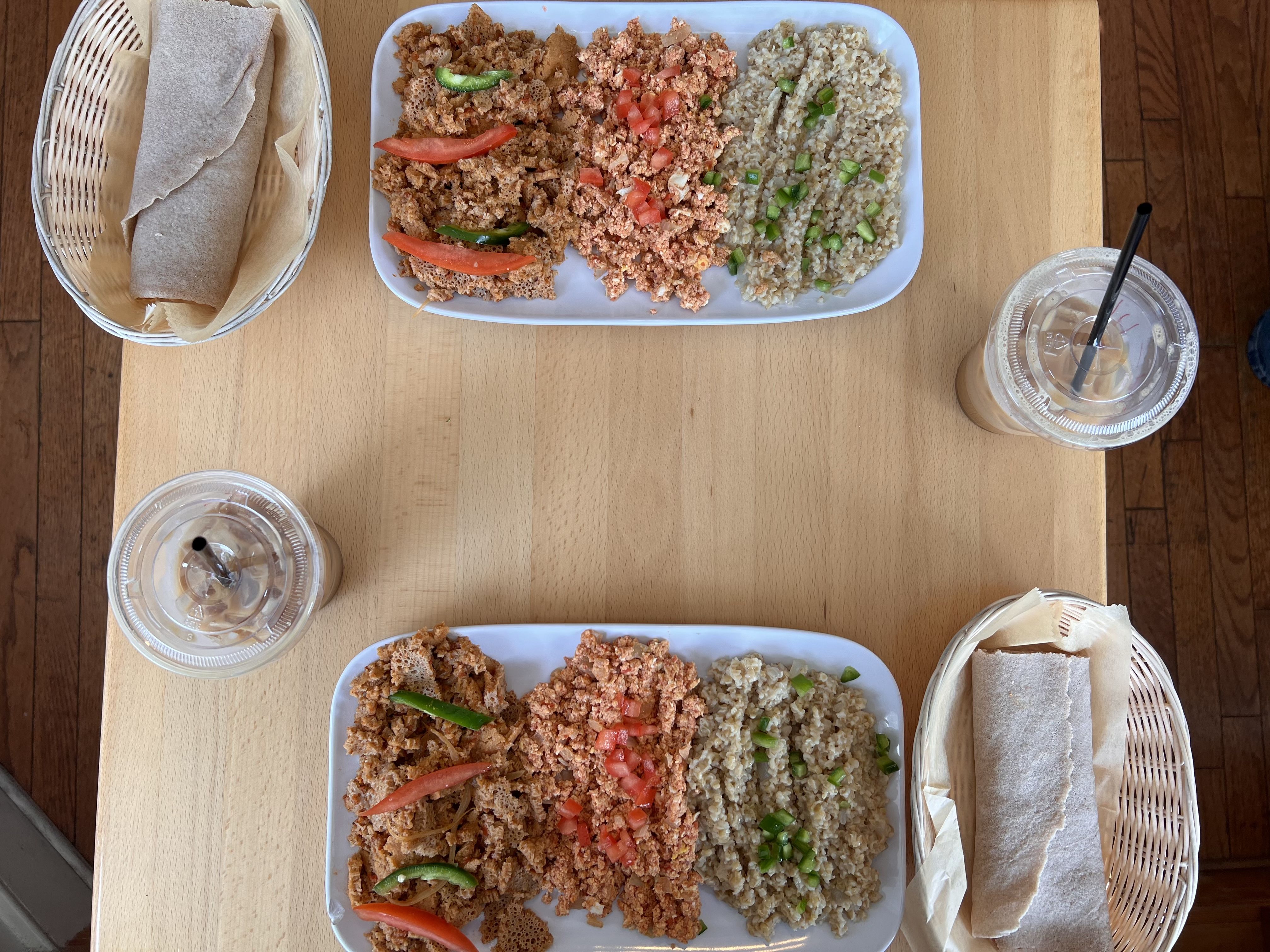 A wooden table with two iced lattes in plastic cups and two trays of food.