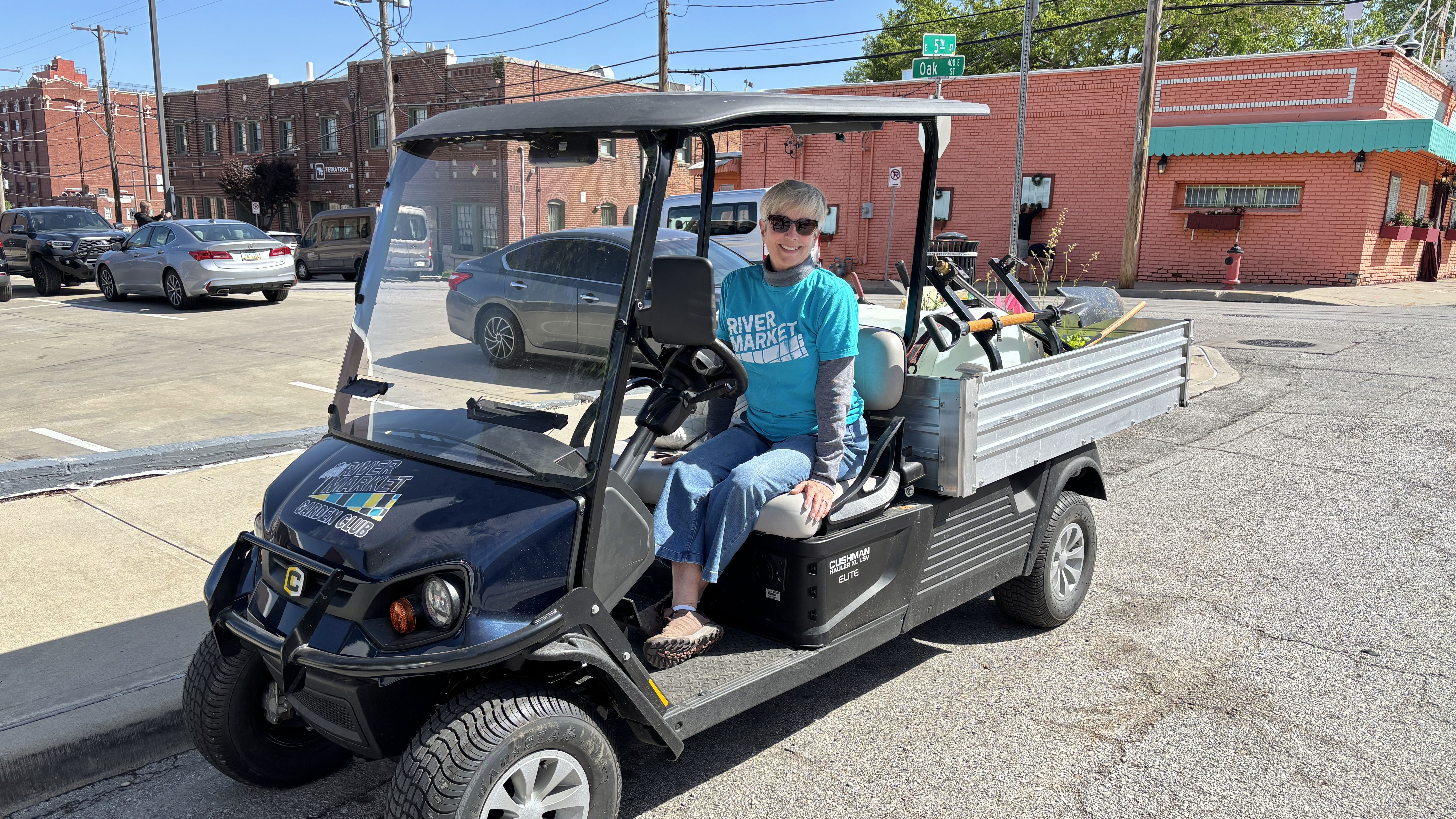 Smiling woman in a turquoise River Market t-shirt sits in a small utility golf cart on a sunny urban street; brick storefronts, parked cars, and a load of tools in the cart's bed.