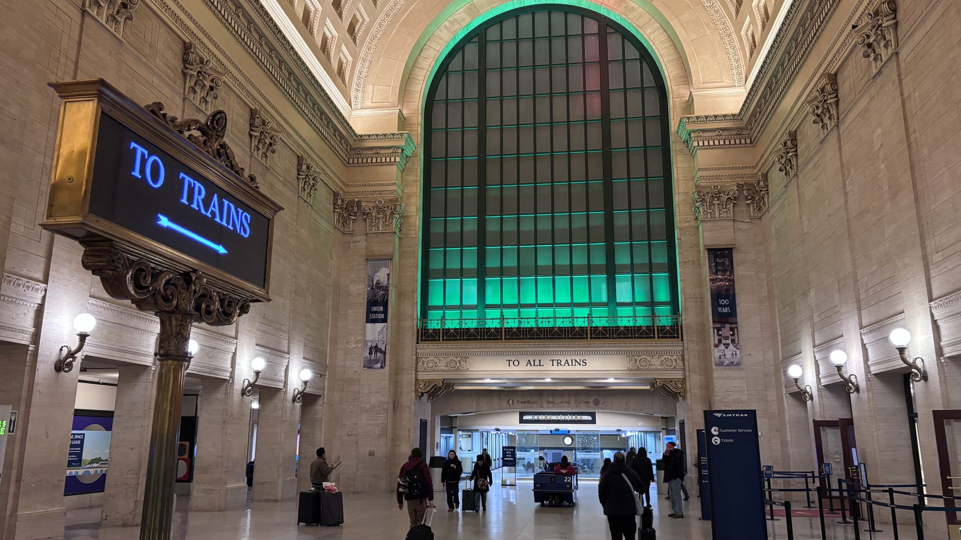 Interior of a large train station with beige walls, a tall arched window lit green, and a blue lit sign reading "TO TRAINS" guiding travelers pulling suitcases toward platforms.