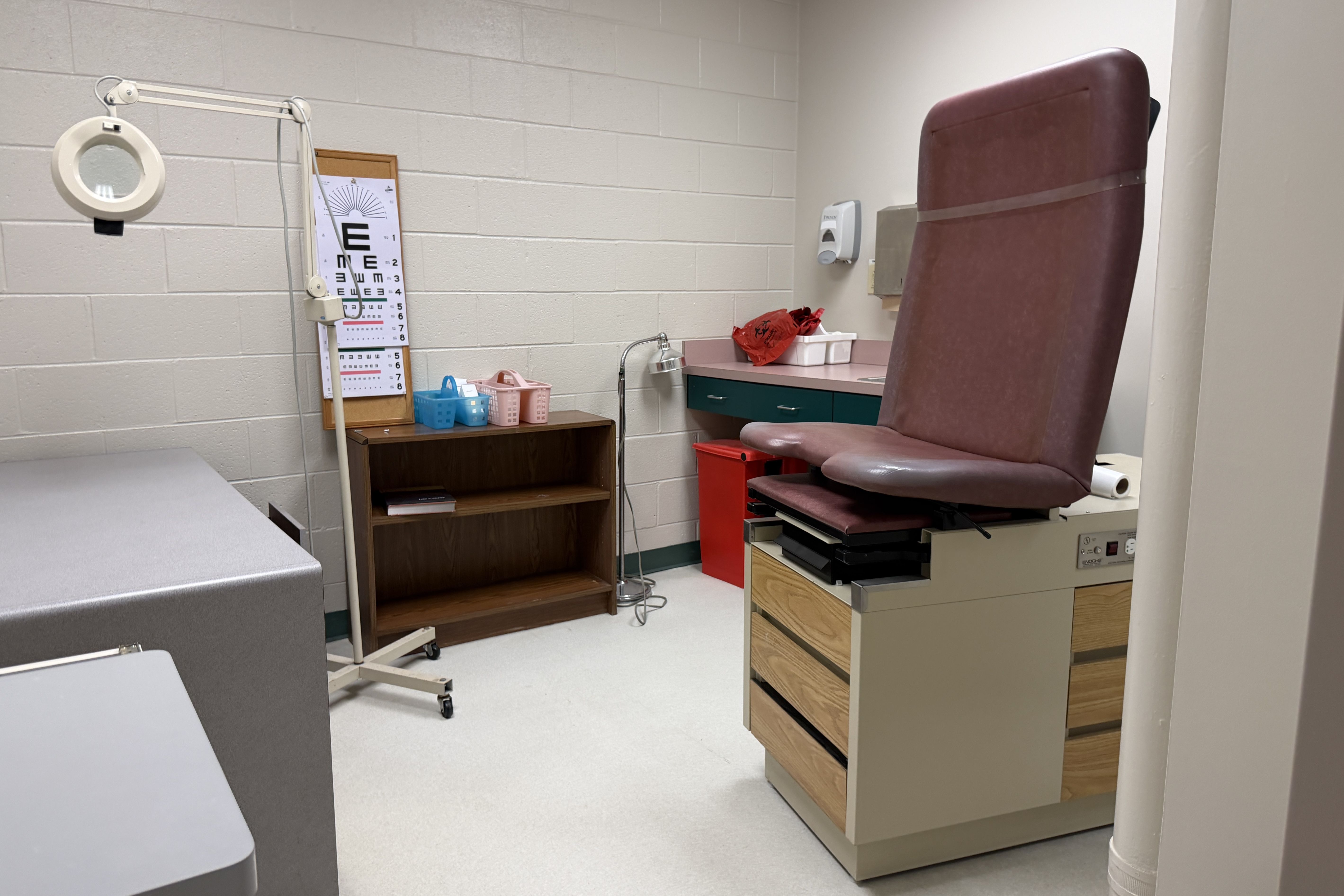 Medical exam room featuring a large burgundy patient chair on the right, Snellen eye chart on the wall, a circular magnifier device, a sink, and assorted medical supplies and carts.