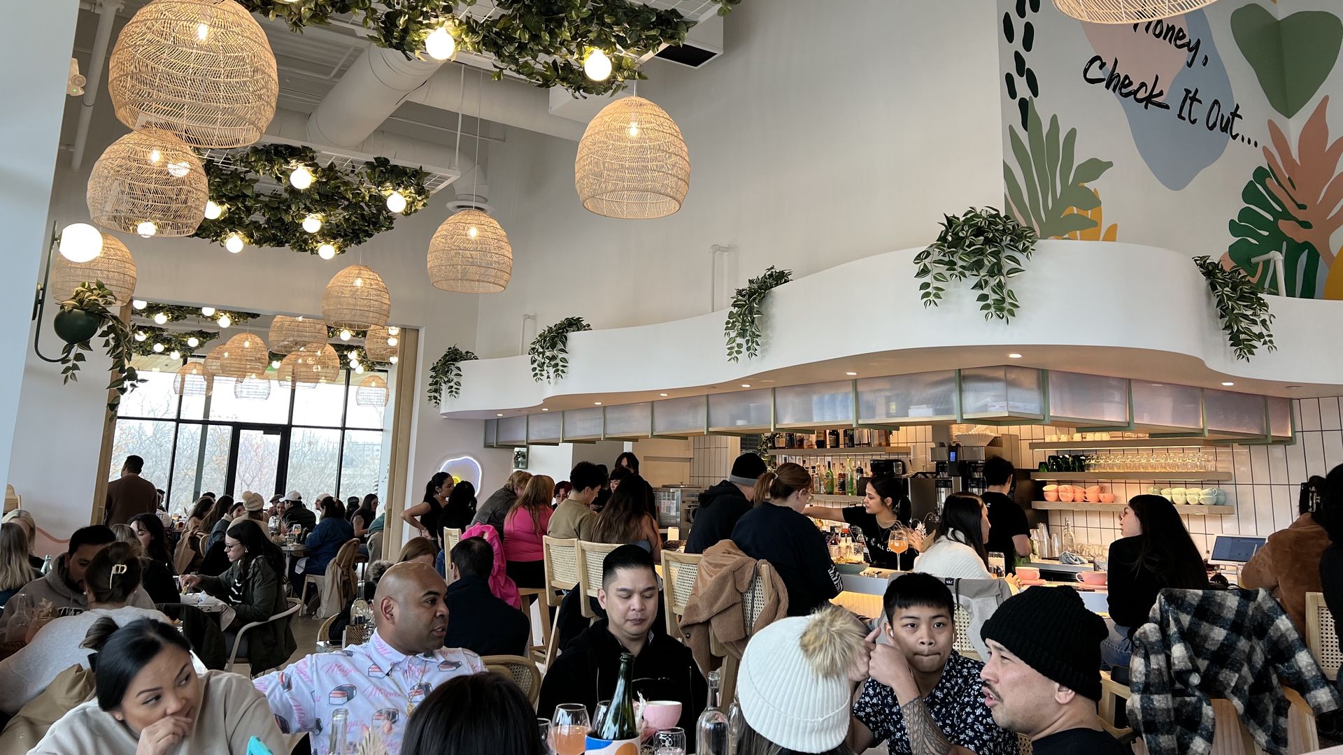 A busy modern cafe with wicker lamps and greenery hanging from the ceiling, white walls, a colorful mural reading "Honey, check it out...", and diverse patrons seated at tables and the bar.