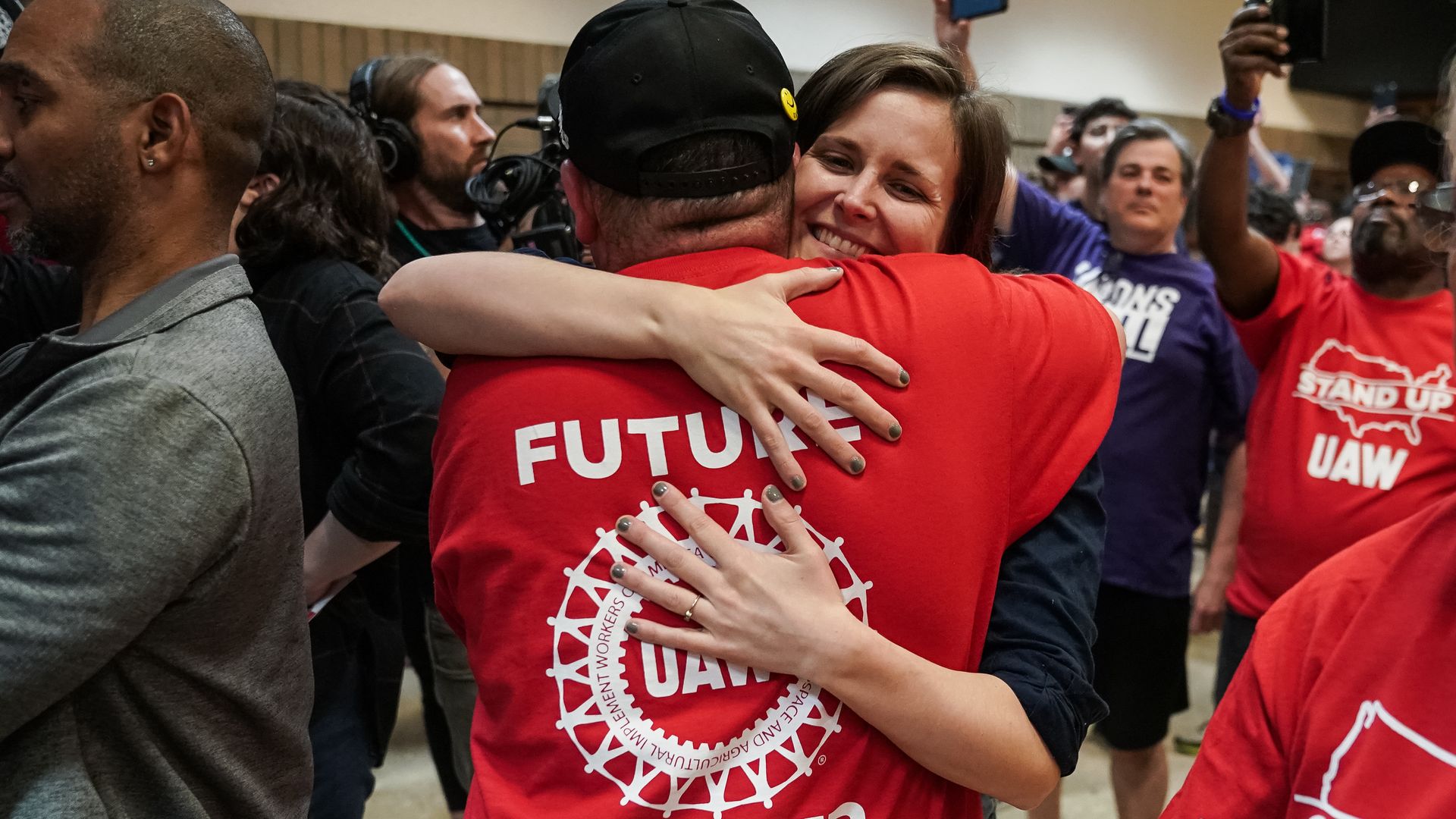 A person in a red shirt hugs another person while others hold cameras