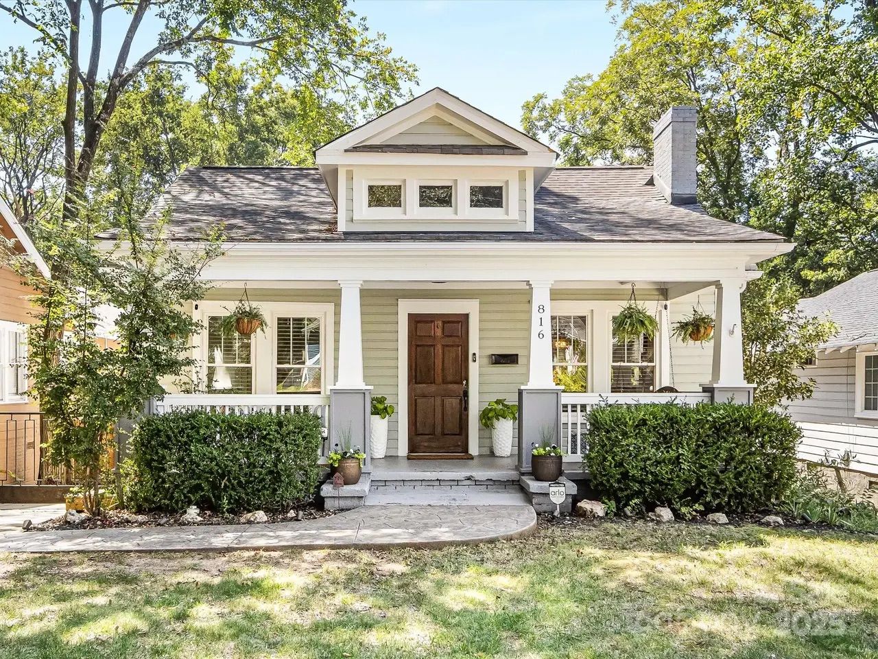 Light gray house with white trim, dark wood front door, porch with white pillars, hanging plants, green bushes, sidewalks, and trees in the background on a sunny day.