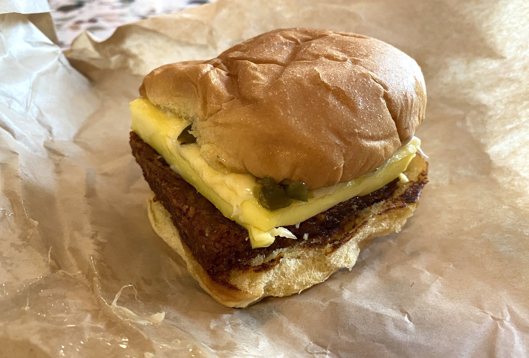 Close-up of a cheeseburger on parchment: a toasted bun, beef patty, melted cheddar cheese, and pickles visible beneath the cheese.