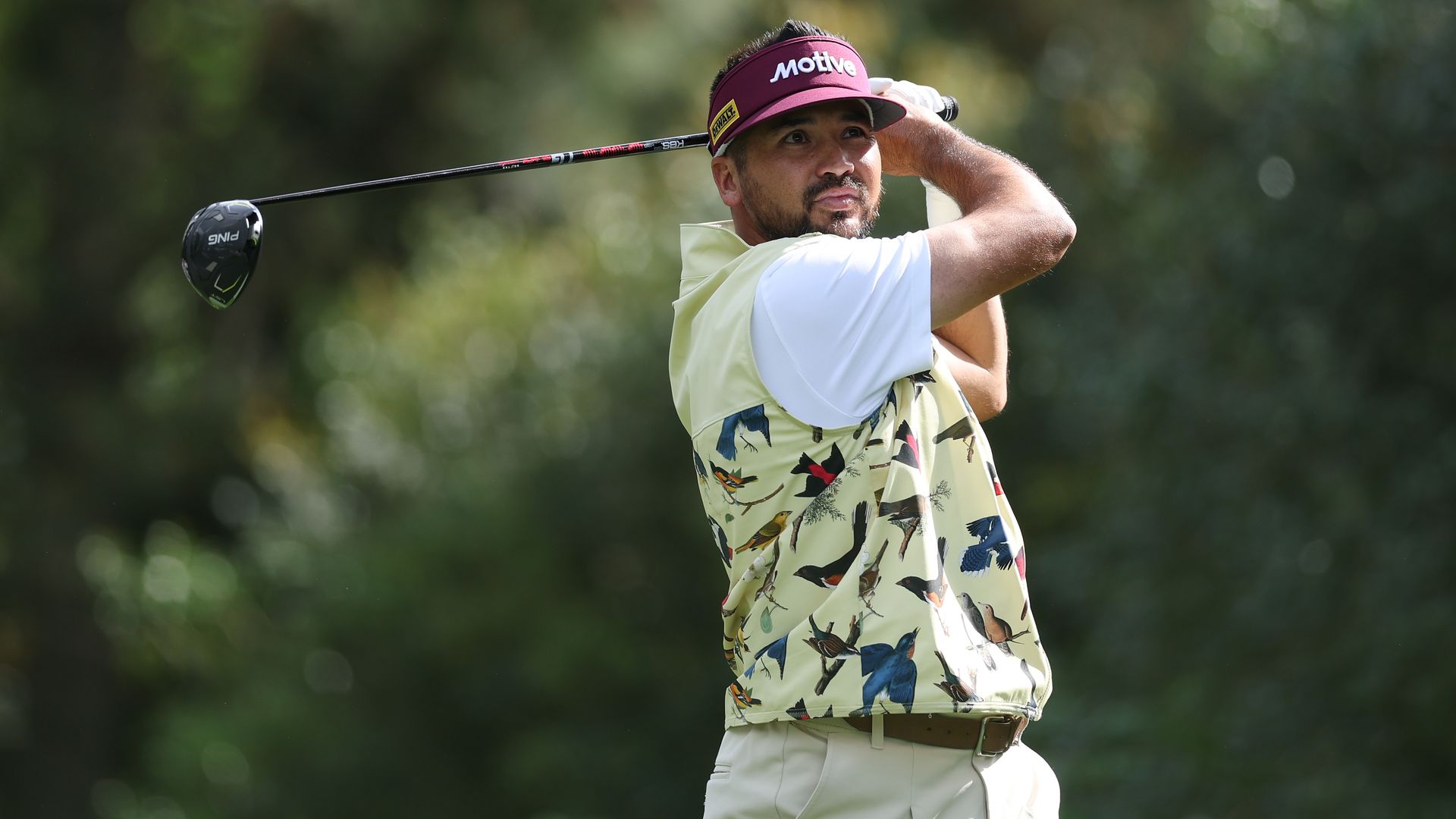 Golfer Jason Day in a pale vest with bird prints and a maroon visor swings a driver on a sunlit green course, with blurred trees in the background.