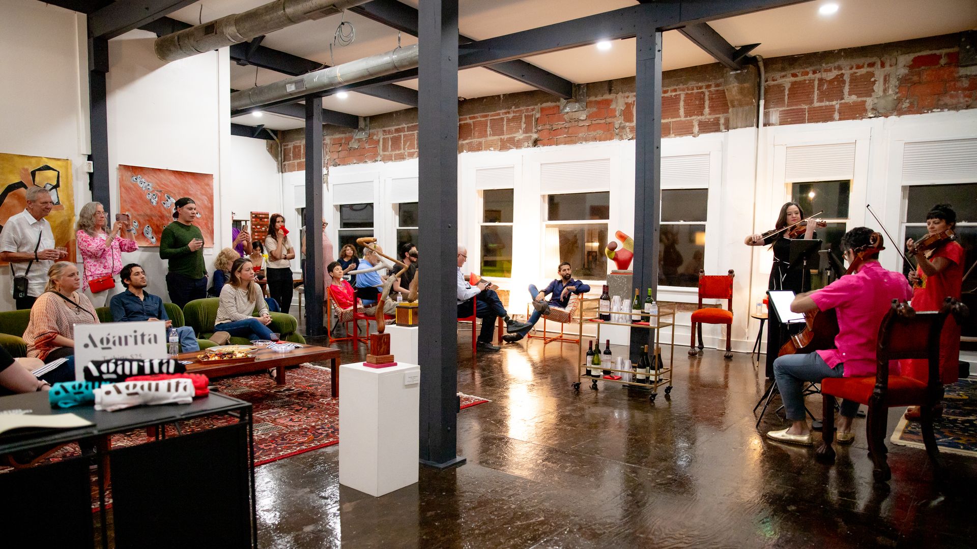 A group of people watch musicians perform in an open space with tall ceilings, metal beams, brick accents and a concrete floor