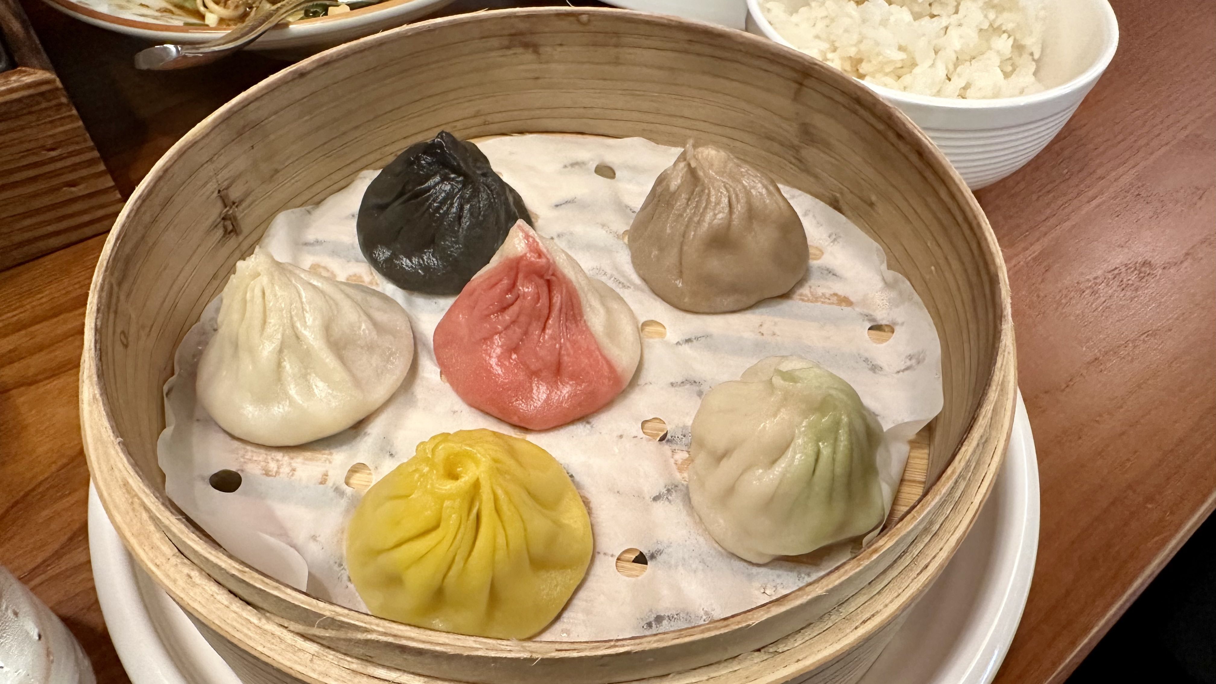 Round bamboo steamer contains several colorful dumplings on parchment; colors include white, black, pink, beige, yellow and green. A bowl of white rice is visible in the background.