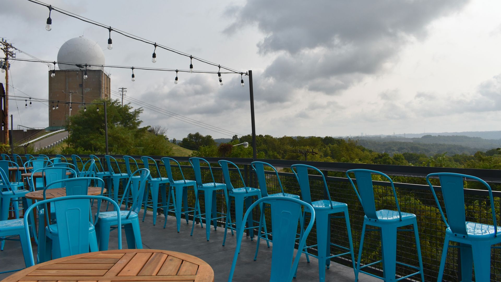 Outdoor terrace with blue metal chairs and wooden tables under string lights, overlooking green trees and a large radar dome with a cloudy sky in the background.