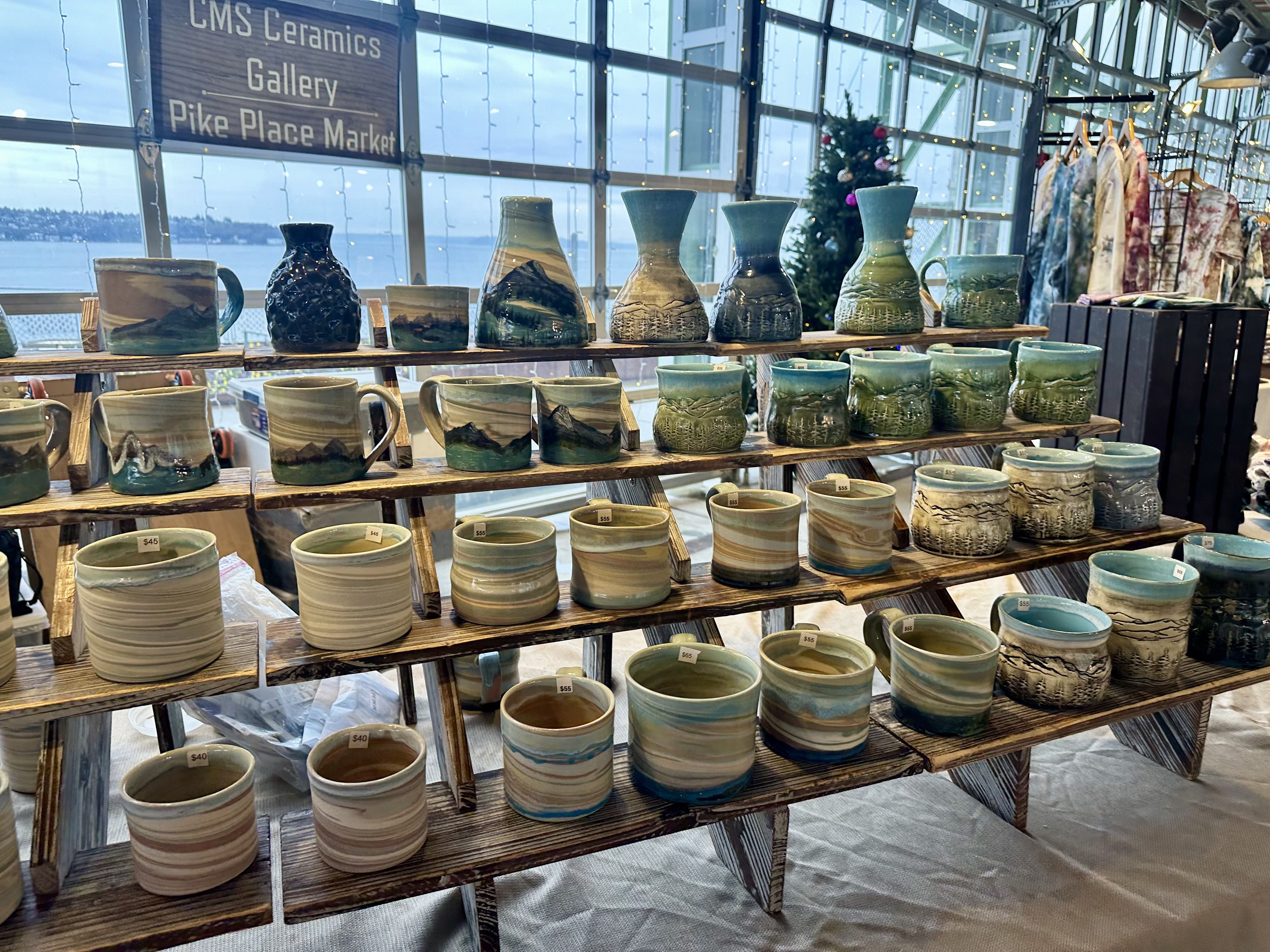 A display of mugs vases and more in earth colors and blues, with roll up garage style windows behind them.