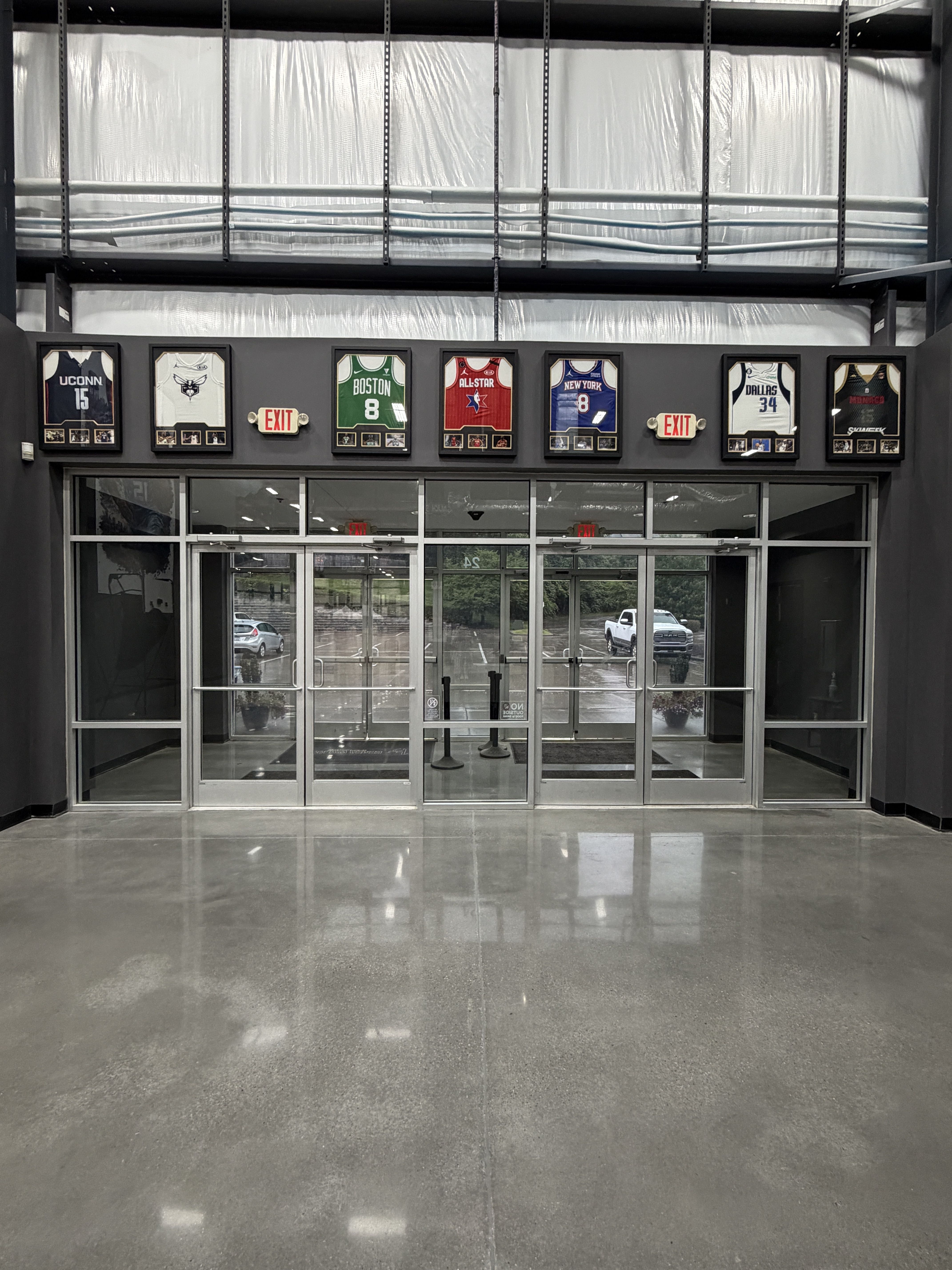 Indoor hallway with framed basketball jerseys on a dark wall: UConn 15, Boston 8, All-Star red, New York 8, Dallas 34, and others; exit signs between frames; glass doors below.