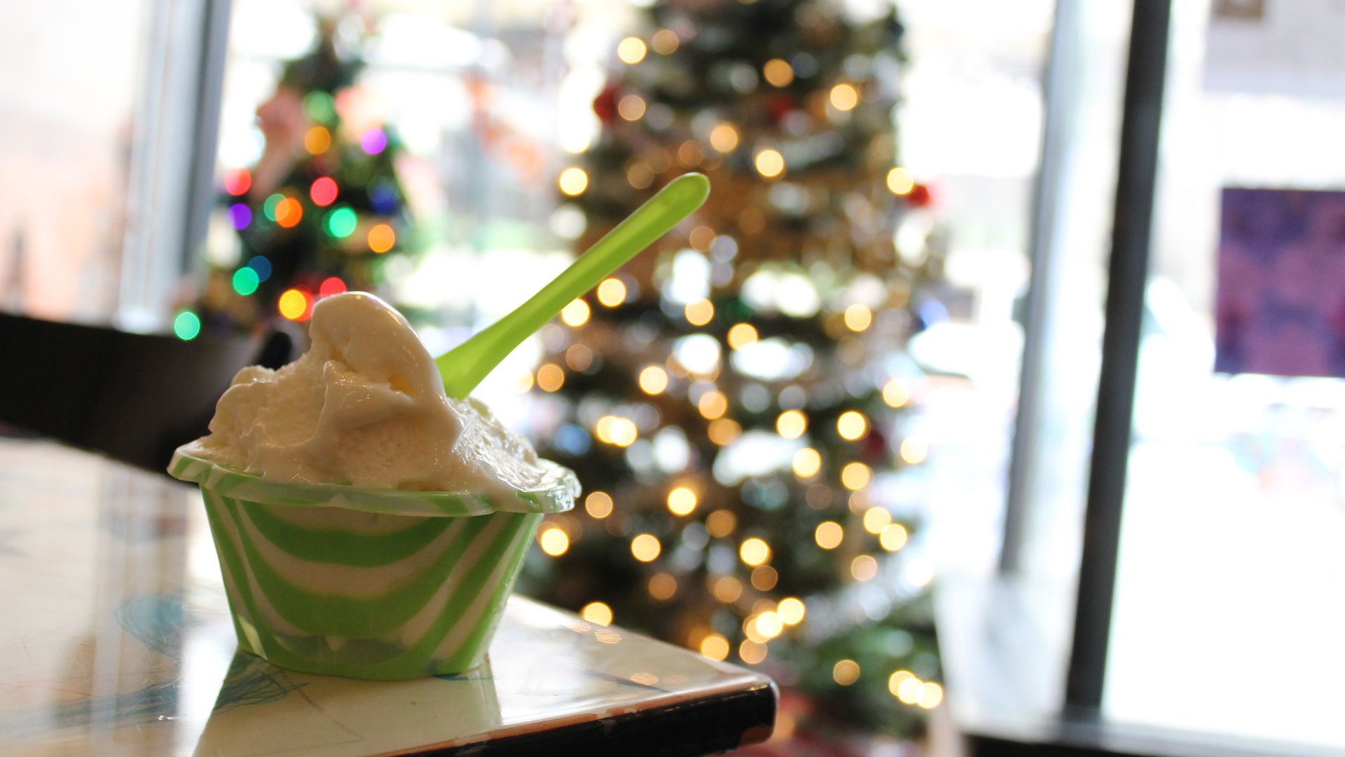 Green and white striped cup of vanilla ice cream with a green spoon on a table, blurry background of decorated Christmas tree with colorful lights.