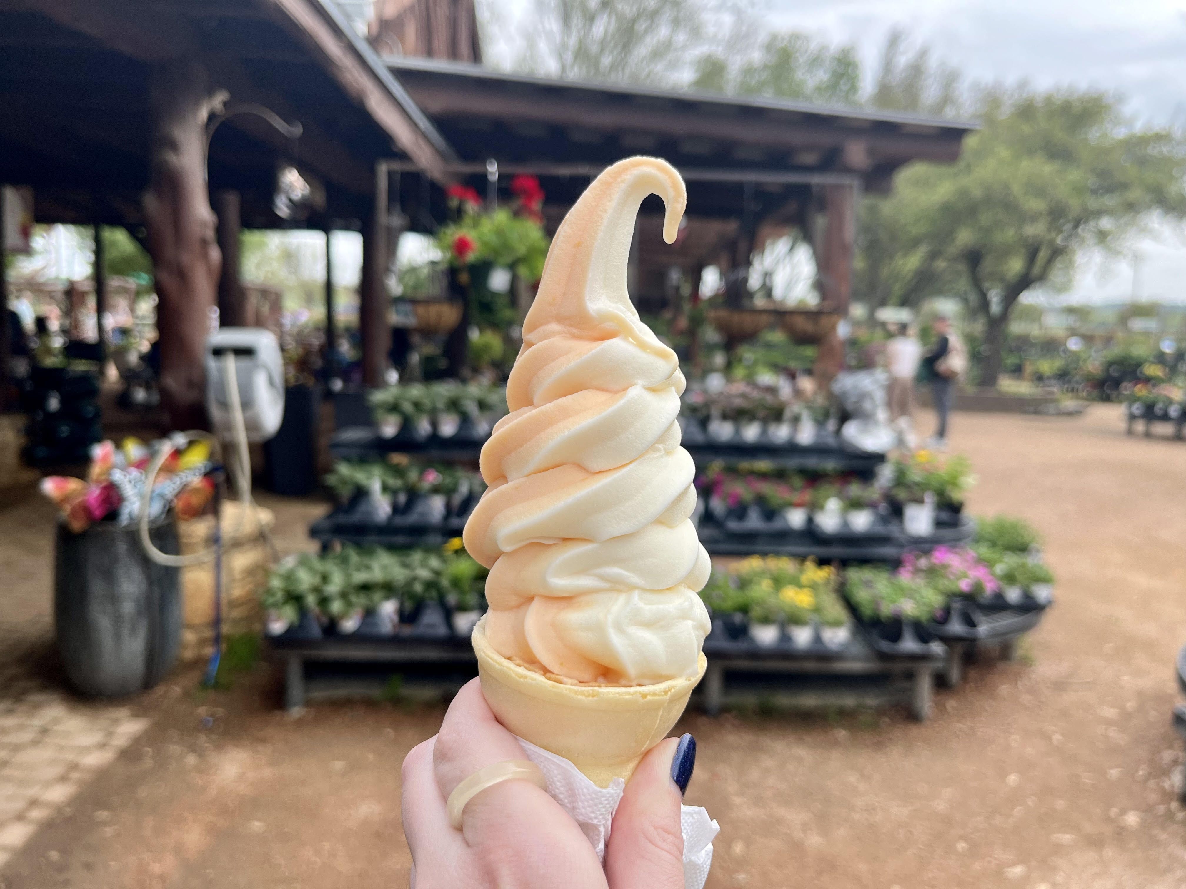 A tall ice scream swirl in a cone with blurred flowers for sale in the background.
