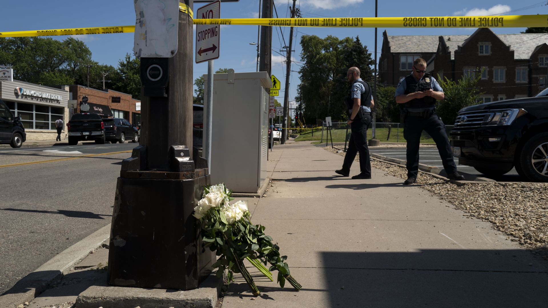 White flowers placed at the base of a black street pole on a sidewalk, yellow police tape reading "POLICE LINE DO NOT CROSS" blocking the area, two police officers in blue uniforms nearby.