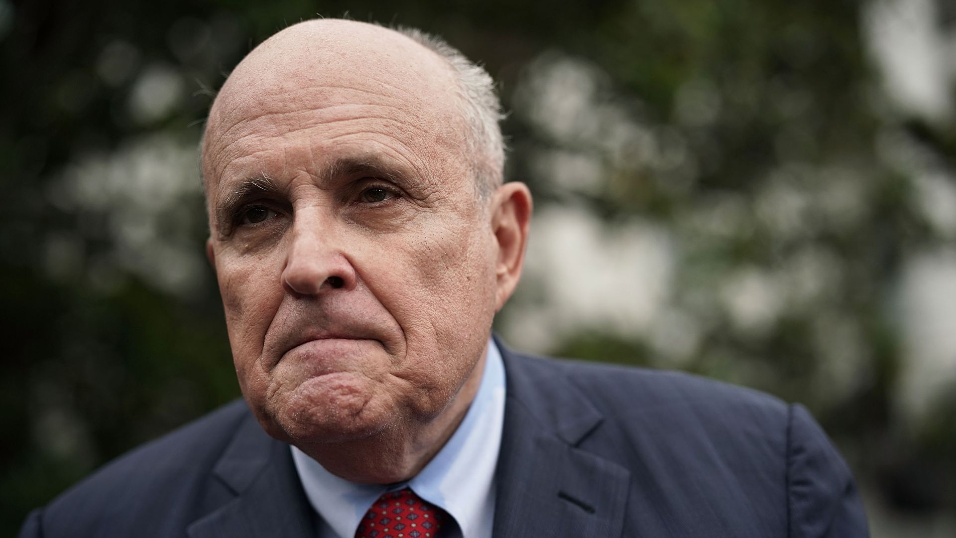 In this image, Rudy Giuliani frowns and walks outside, wearing a suit. 