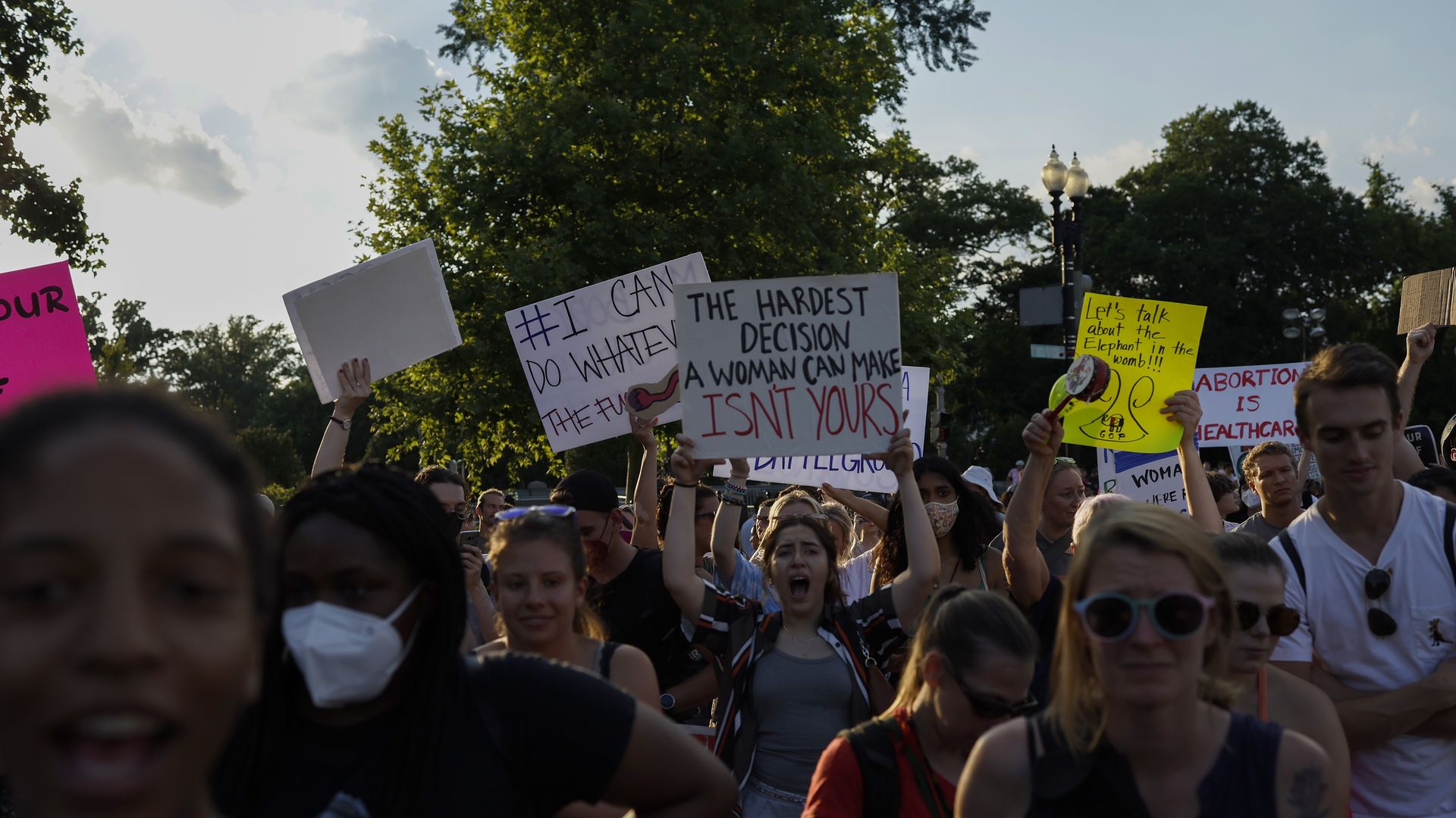 People protest outside the Supreme Court. A crowd of people holding signs is clustered in front of trees. The sign in the middle says: The hardest decision a woman can make isn't yours.