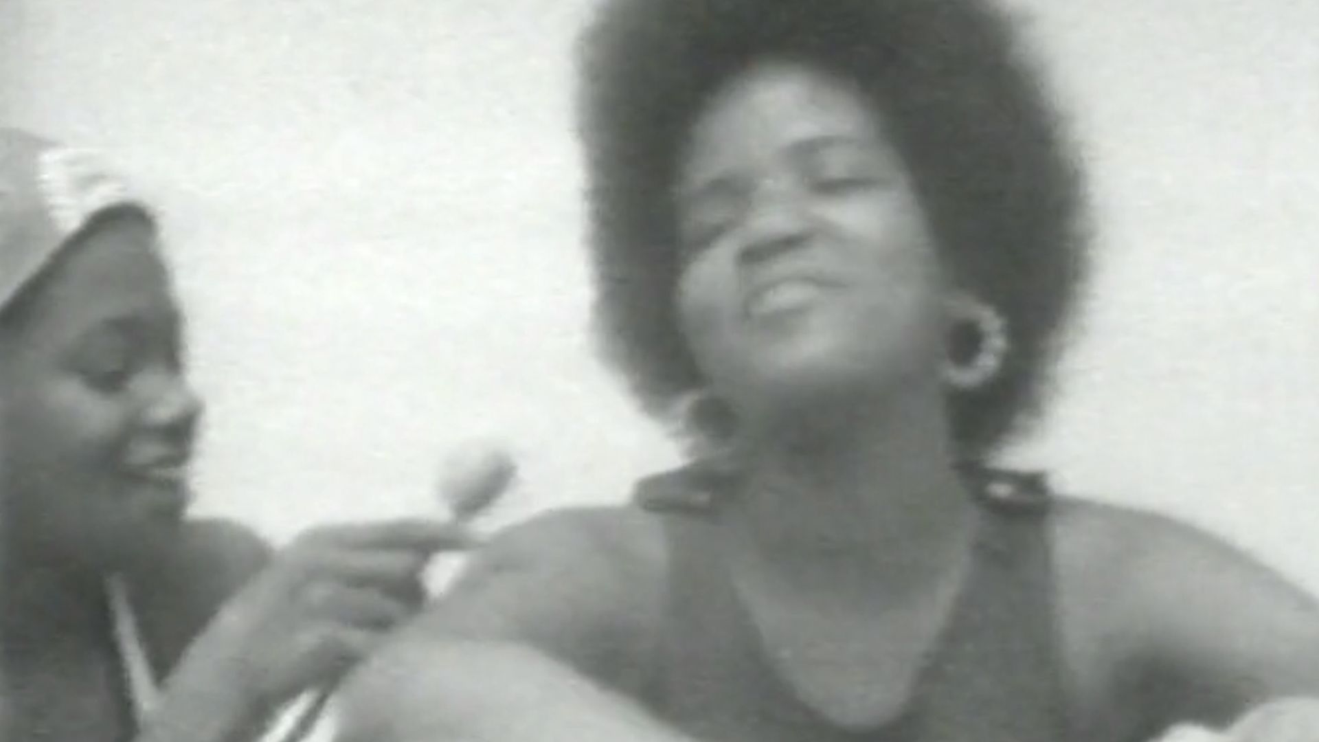 Black and white still of a girl with a microphone interviewing a woman.